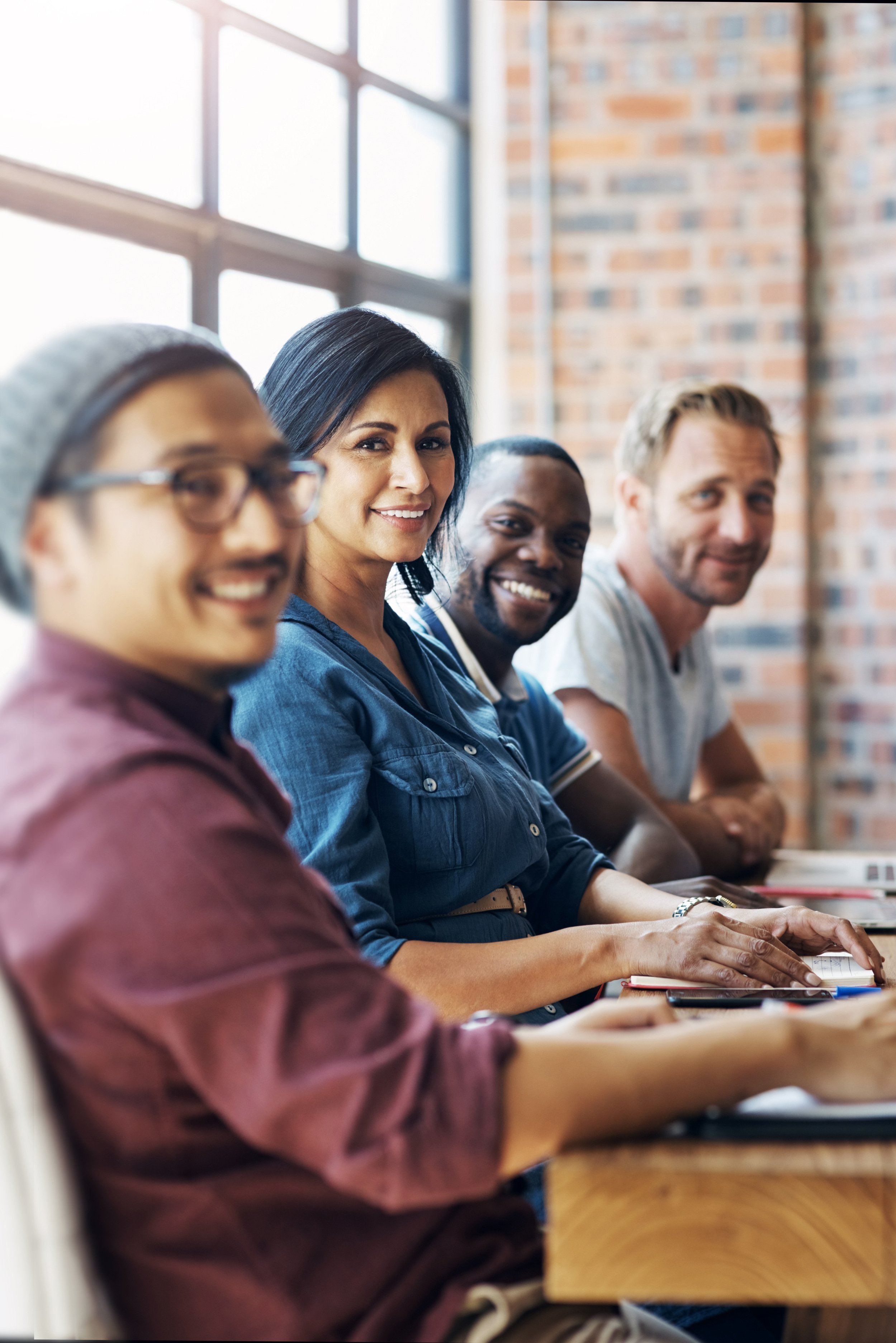 Four ethnically diverse people sitting in a business meeting in a bright room with large windows and exposed brick wall, smiling and looking towards the camera.