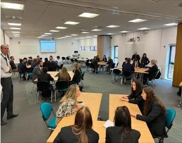 A conference room filled with people seated at tables, with some standing and speaking. A presentation is projected on a screen at the front.