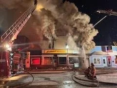 Building on fire with smoke billowing out, firefighters and fire trucks present at the scene, and emergency lights flashing.