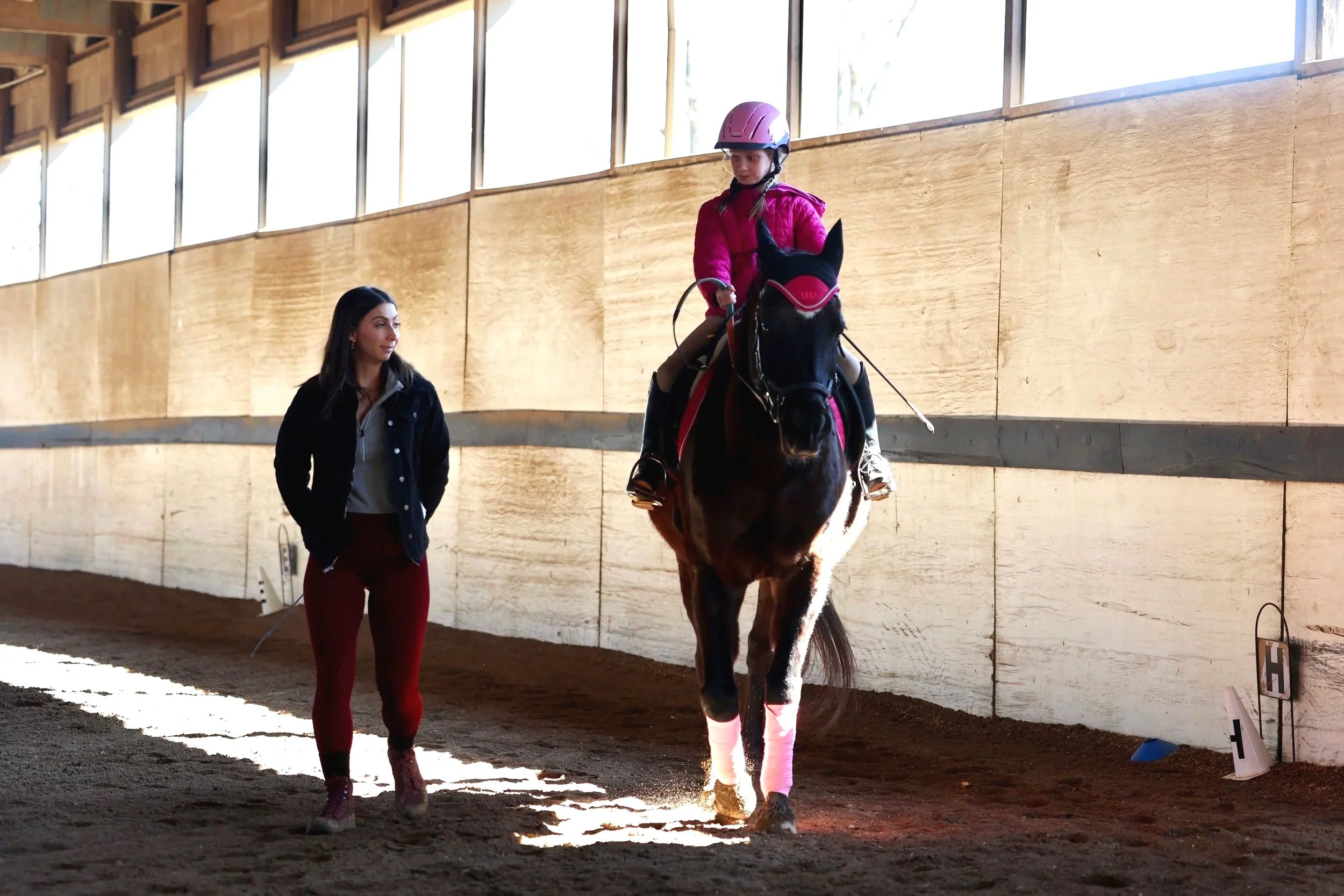A young girl wearing a pink helmet and riding equestrian gear sits on a dark brown horse with pink leg wraps inside an indoor riding arena. A woman in a black jacket and red pants walks nearby, observing the girl.