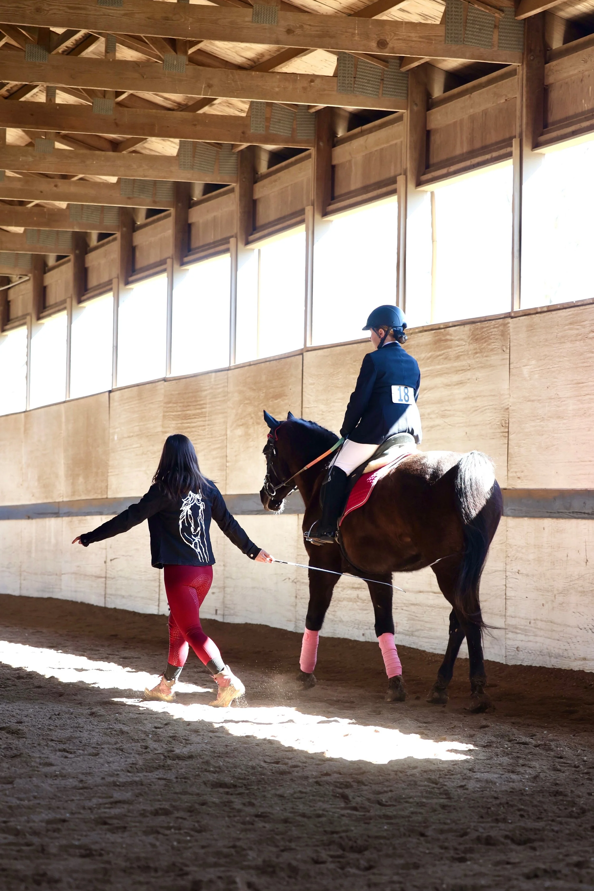 A young rider sitting on a horse inside an indoor riding arena, with a guide walking beside them holding the horse's lead. The rider is wearing a helmet and a black jacket, and the horse has leg wraps on its front legs.