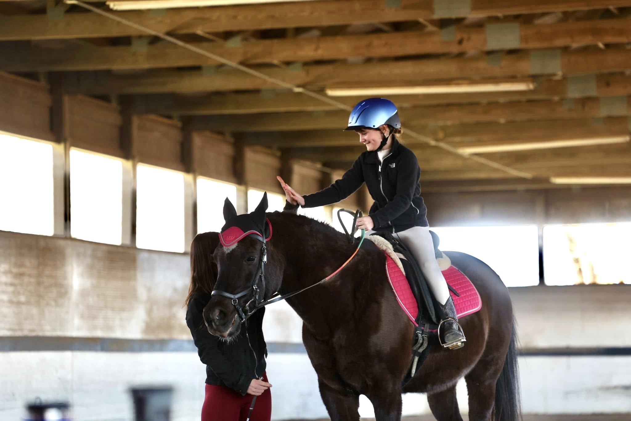 A girl in a riding helmet high-fiving another woman while sitting on a brown horse with a pink saddle and bridle inside an indoor riding arena.