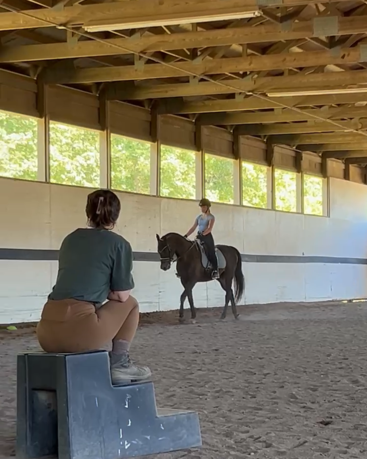 A person sitting on a low platform watches a girl riding a horse inside an indoor riding arena with large windows letting in natural light.