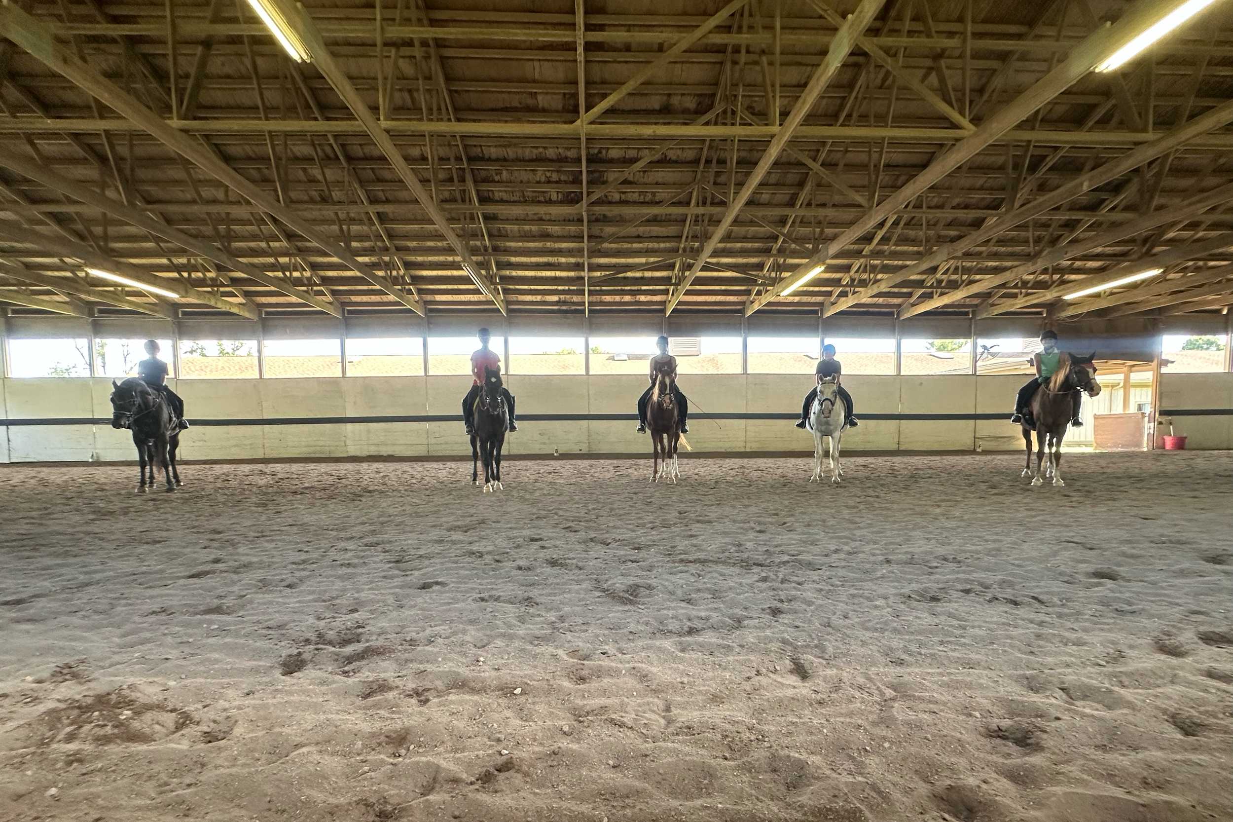 Five people riding horses inside an indoor equestrian arena with a dirt floor and a wooden roof.