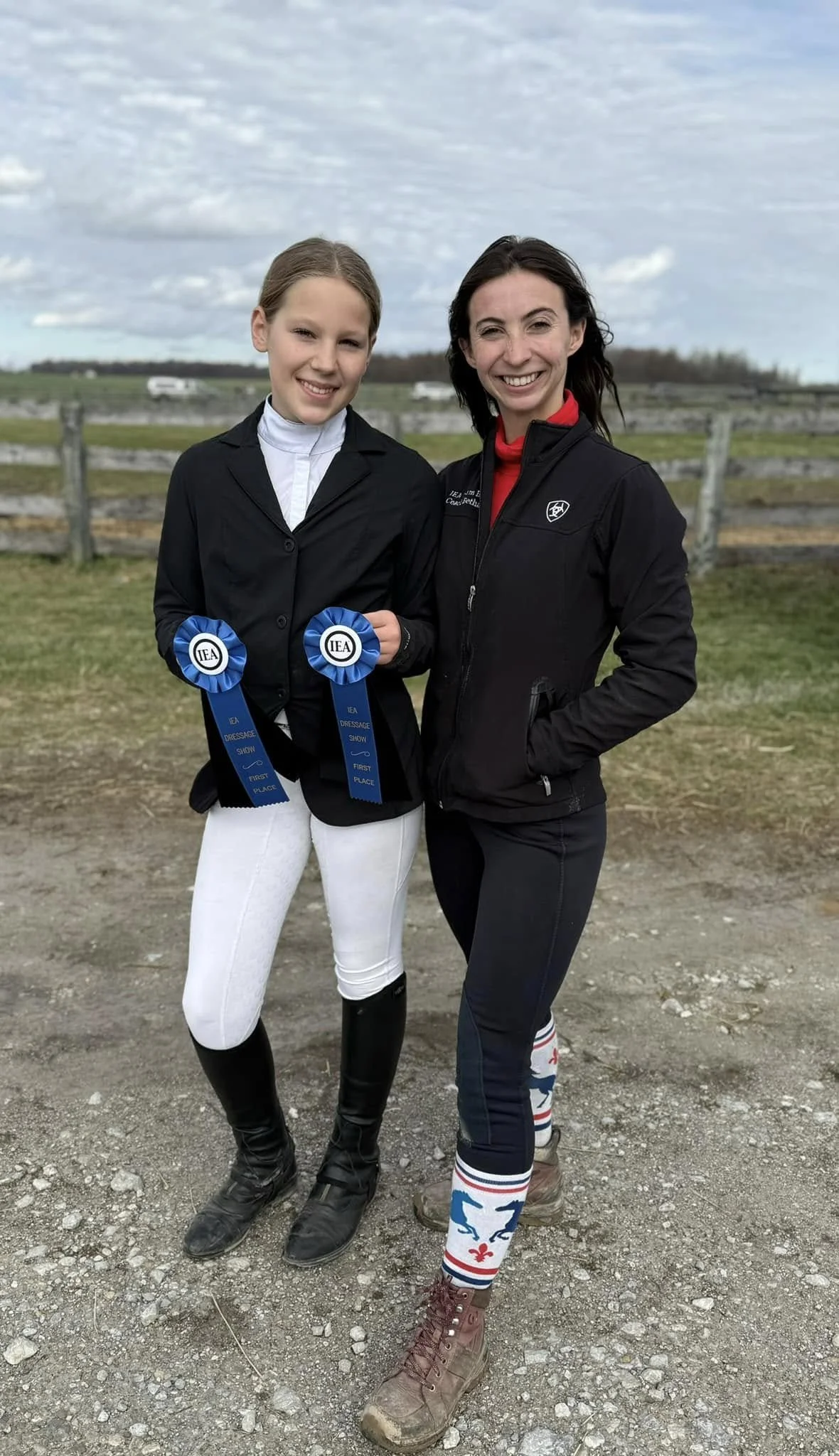 Two women standing outdoors on a cloudy day, one holding two blue and white ribbons for first place in a dressage show, dressed in equestrian attire including riding boots and breeches, the other wearing a black jacket, patterned socks, and casual cl