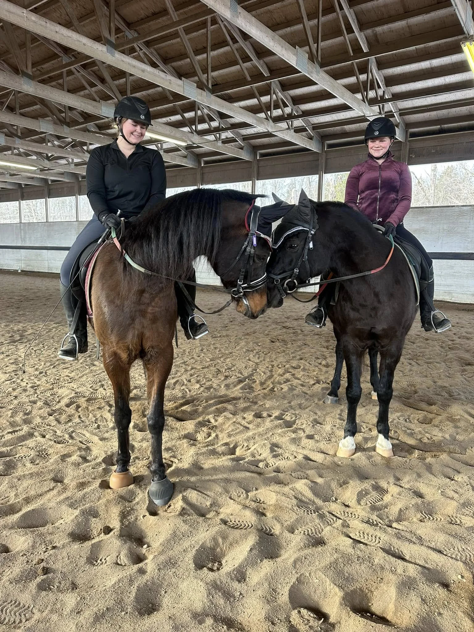 Two girls riding horses in an indoor riding arena, touching heads with the horses, wearing helmets and riding gear, with sandy footing and a wooden ceiling.