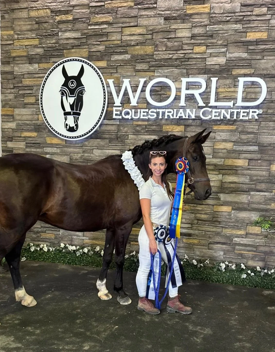 A woman standing next to a horse with a blue and yellow ribbon at the World Equestrian Center. The woman is smiling and has a flower garland around her neck, in front of a stone wall with a sign that says 'World Equestrian Center' and a horse head logo.