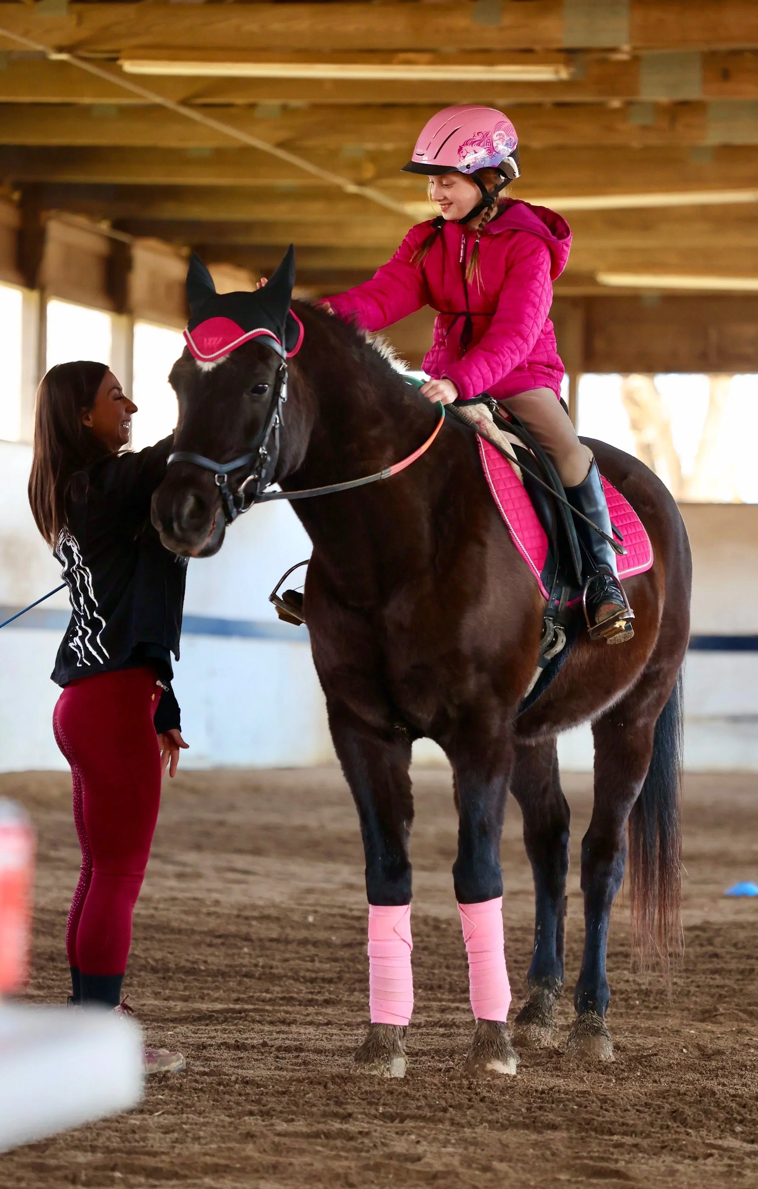 A young girl wearing a pink helmet and jacket riding a brown horse inside an indoor riding arena. An instructor stands nearby, holding the horse's bridle, smiling and engaging with the girl.