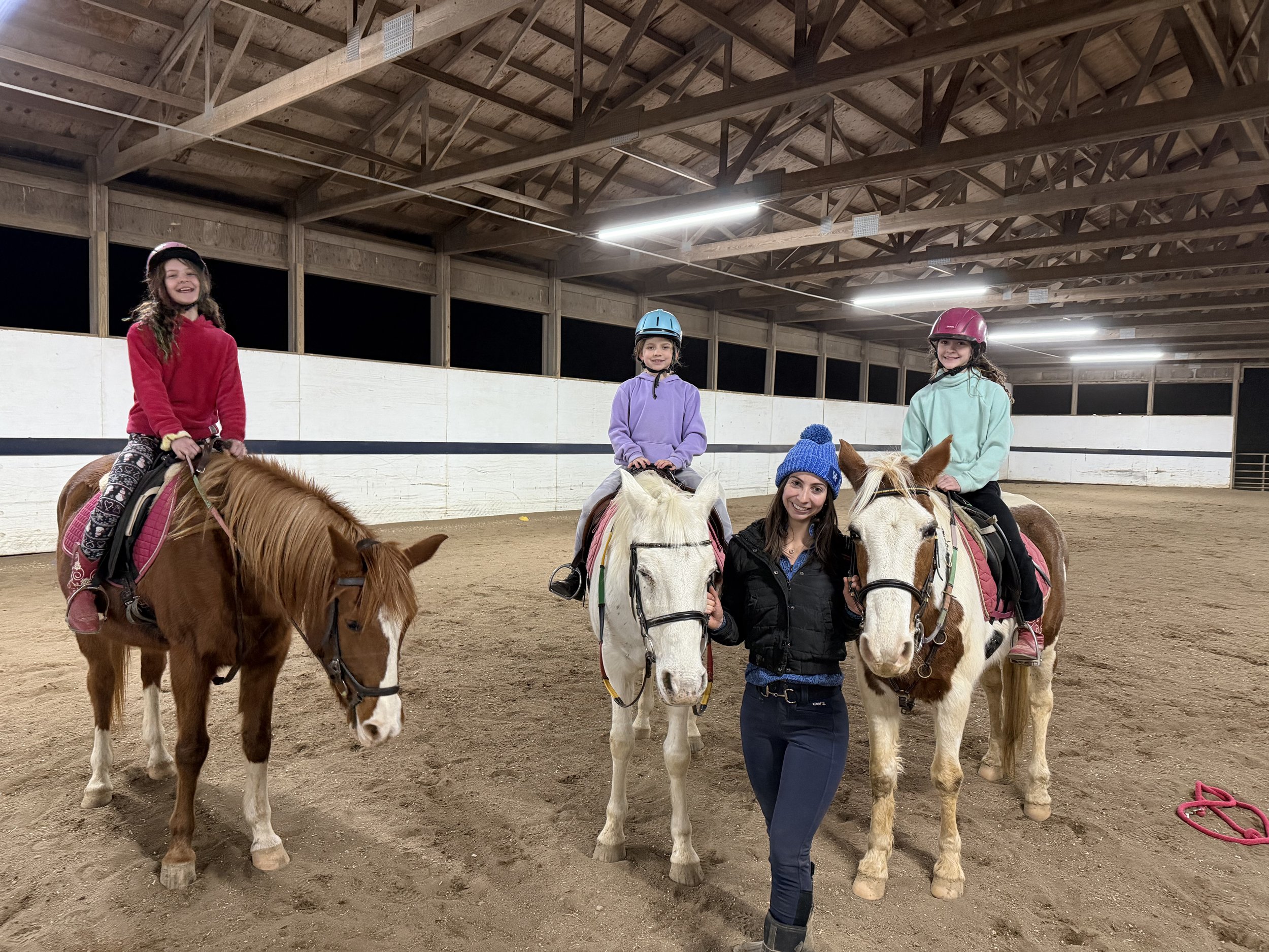 Three young girls riding ponies in an indoor riding arena with a woman standing between the ponies, holding their reins and smiling.