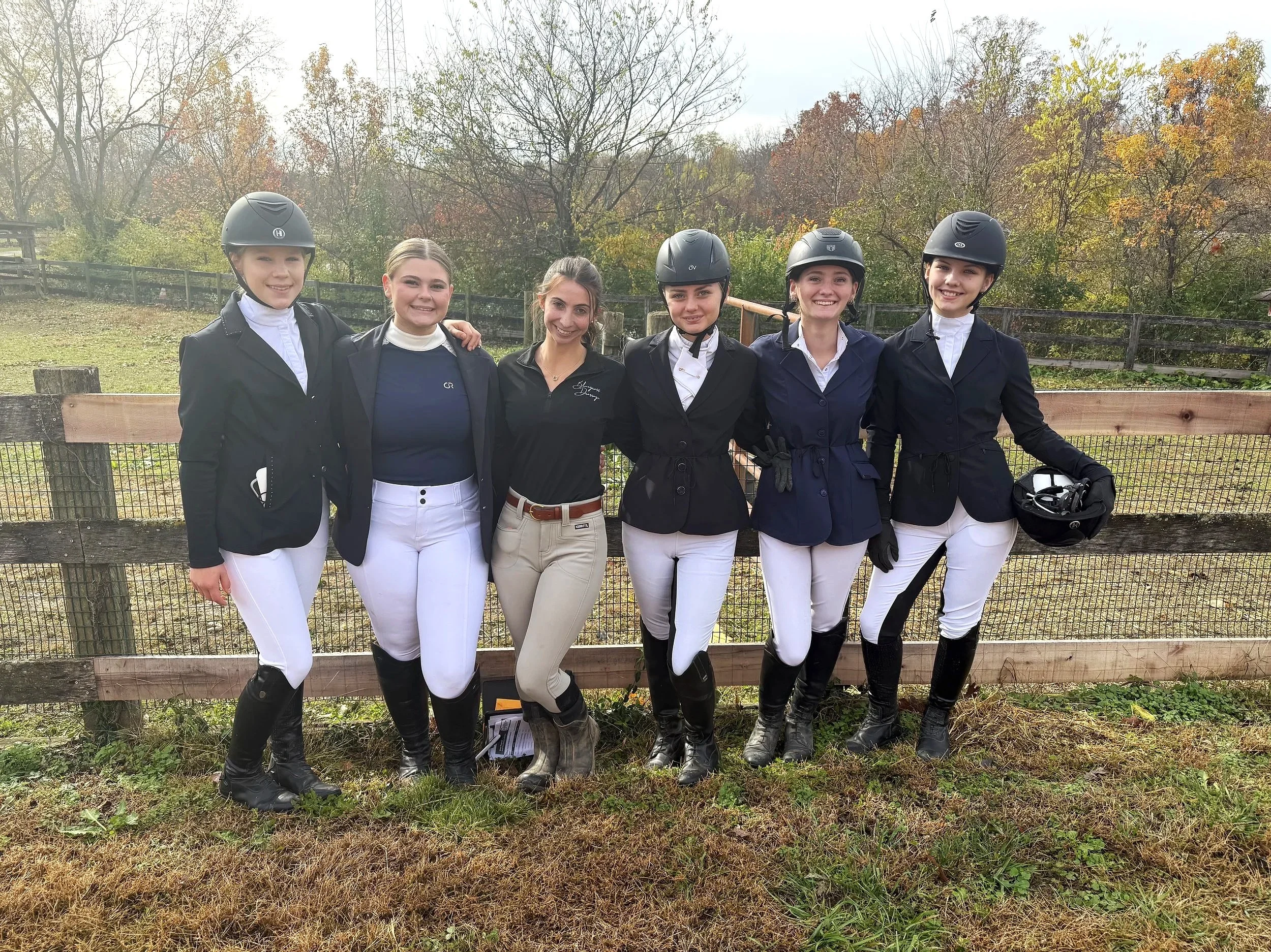 Six young women dressed in riding gear, standing outdoors by a wooden fence, smiling for the camera, with autumn trees in the background.
