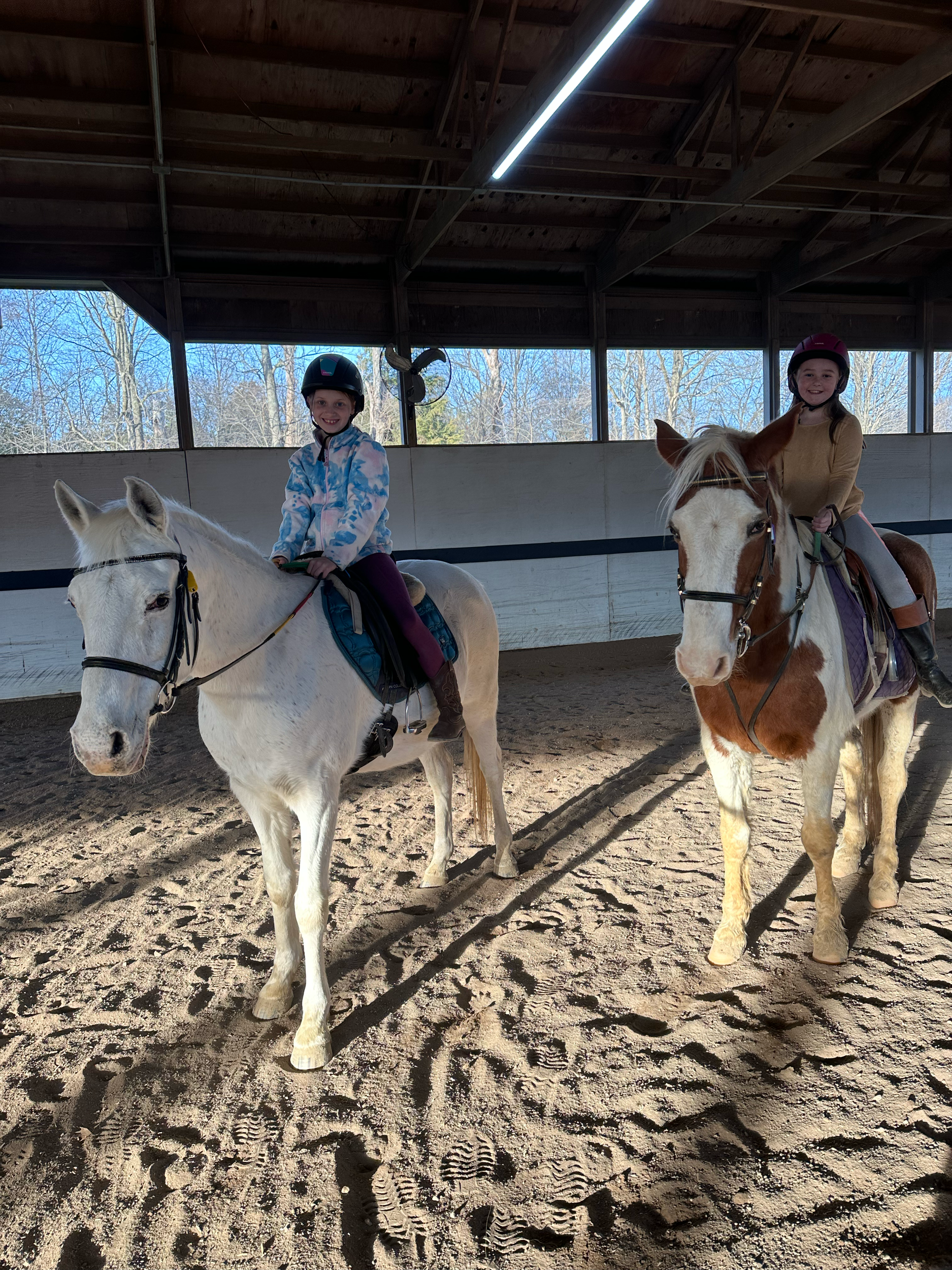 Two children riding white horses inside an indoor riding arena, wearing helmets and riding gear.