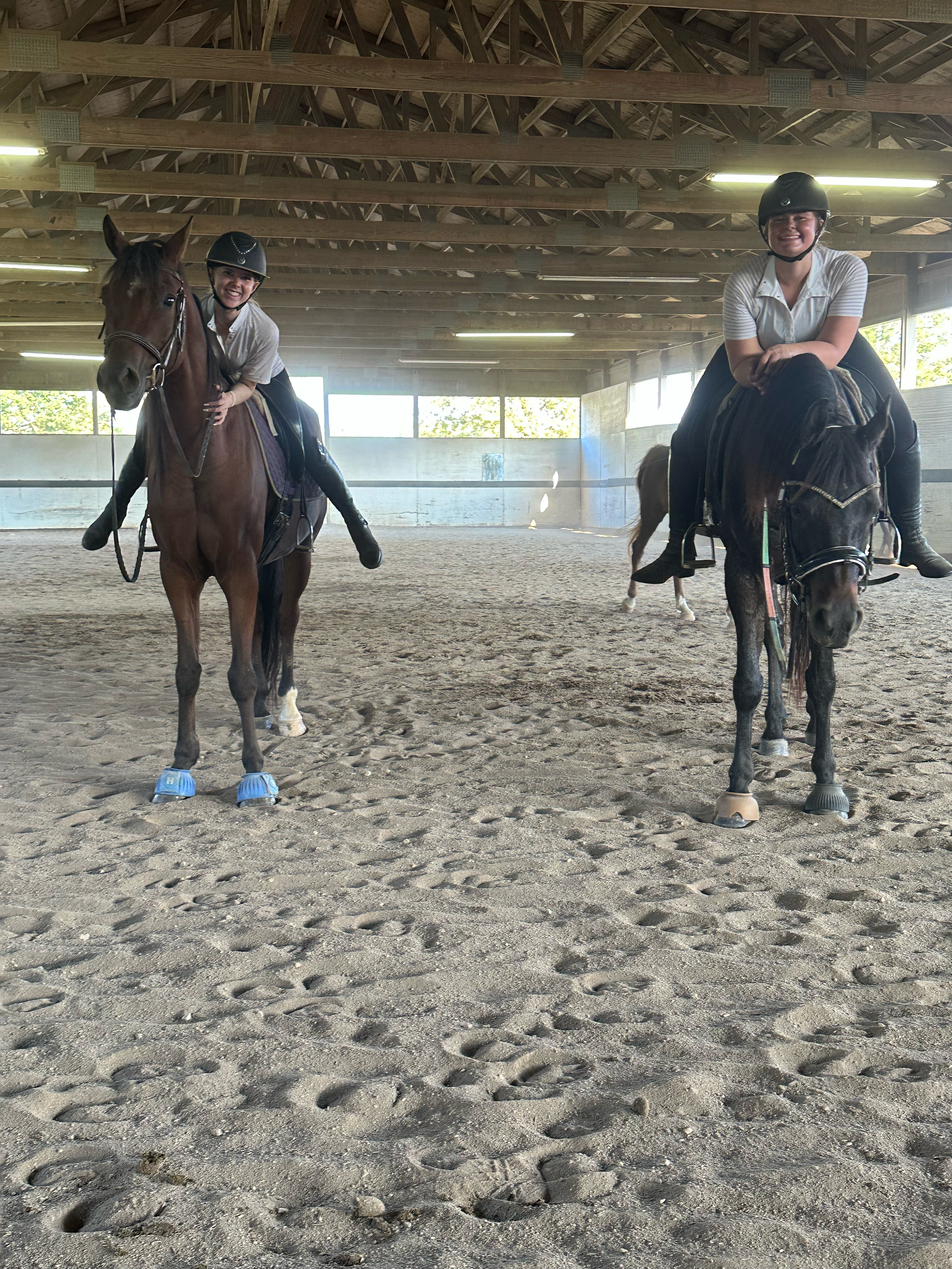 Two women sitting on horses inside a riding arena, wearing helmets and riding gear, with a wooden roof structure and large windows in the background.