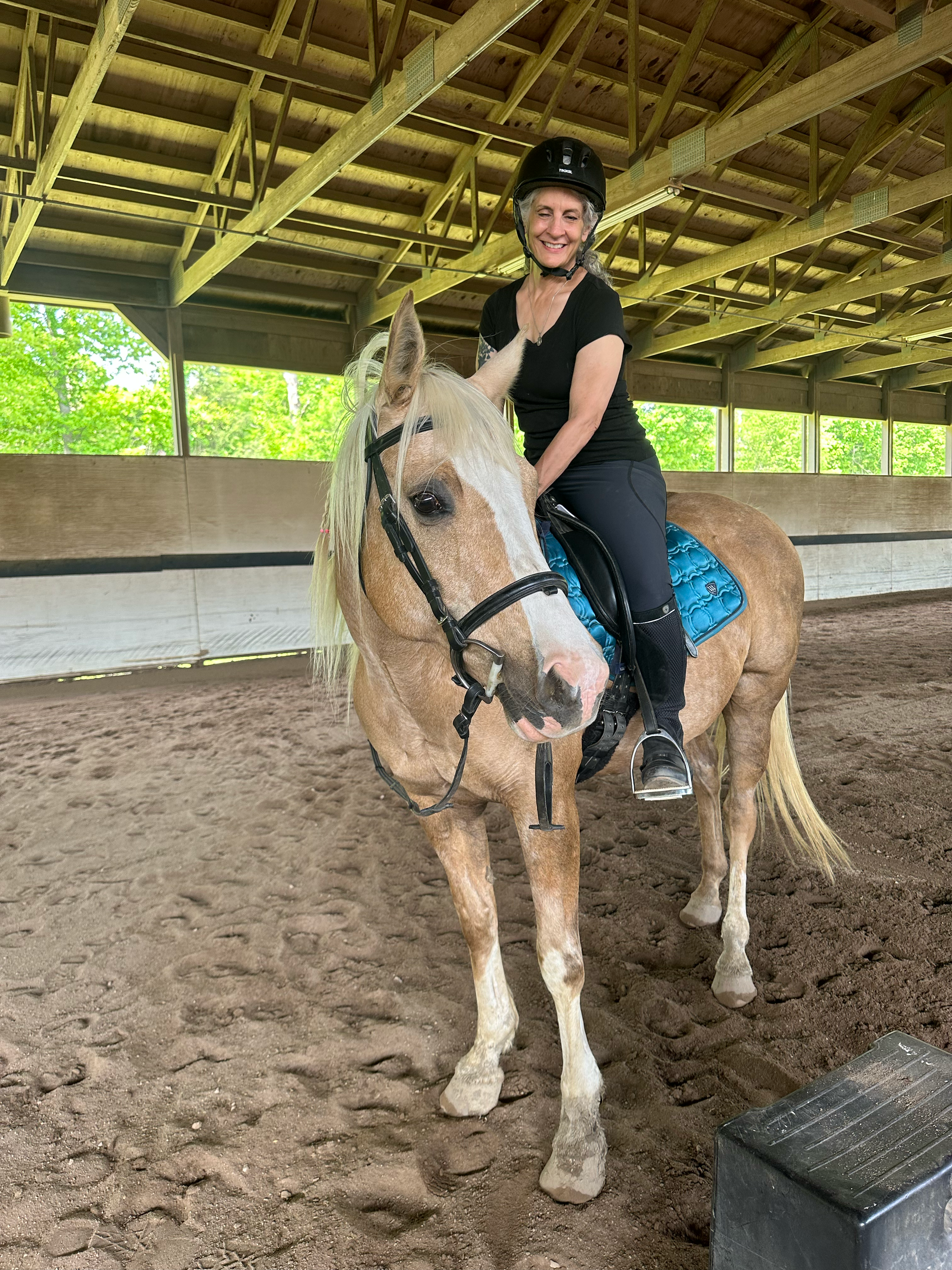 A woman with a black riding helmet, black shirt, and black pants sits on a light tan horse with a white mane inside an indoor riding arena. The horse has a blue saddle pad and black bridle. The arena has wooden structure and large windows with trees 