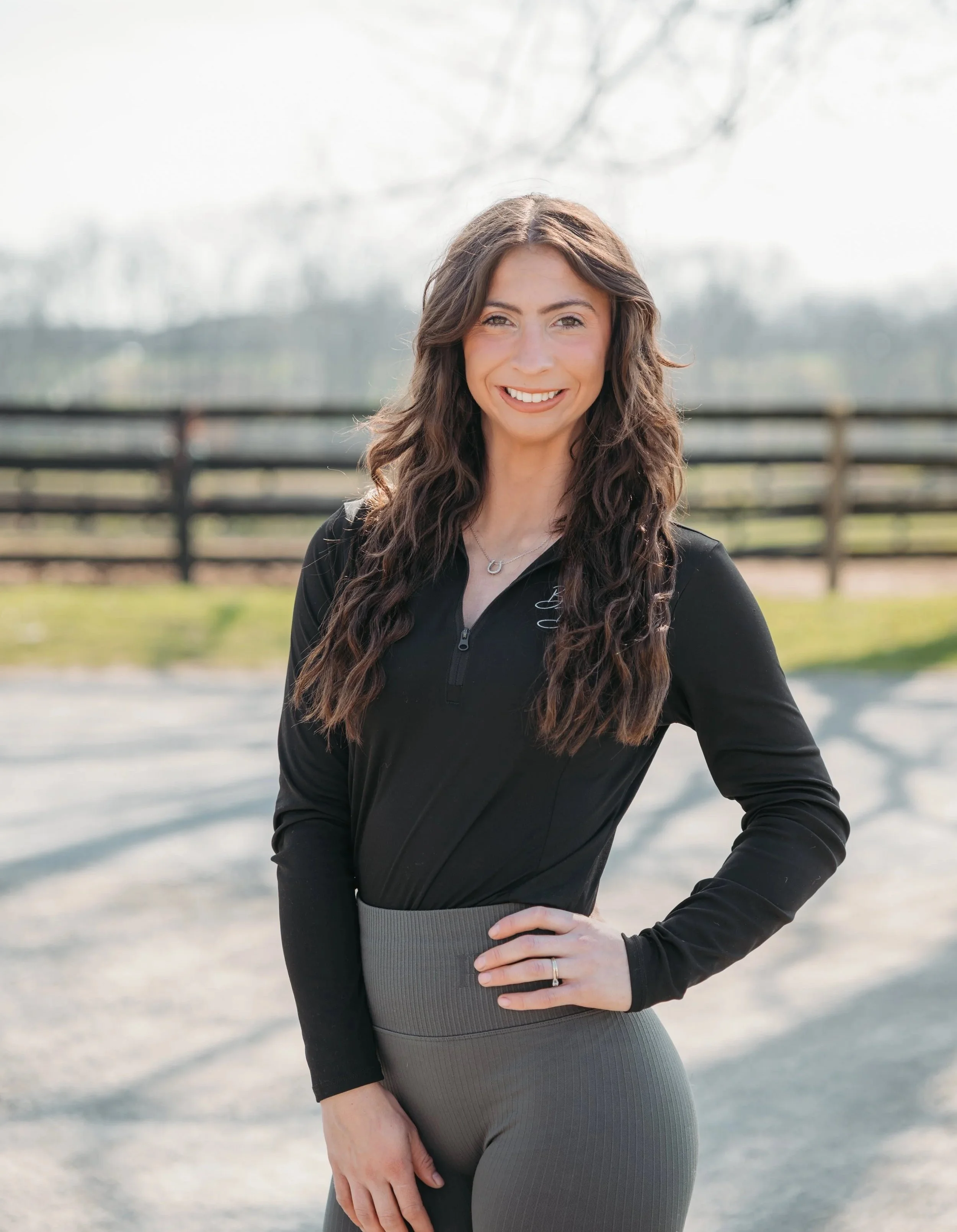 A young woman with long, wavy brown hair, wearing a black zip-up jacket and gray leggings, smiling outdoors with a fence and trees in the background.