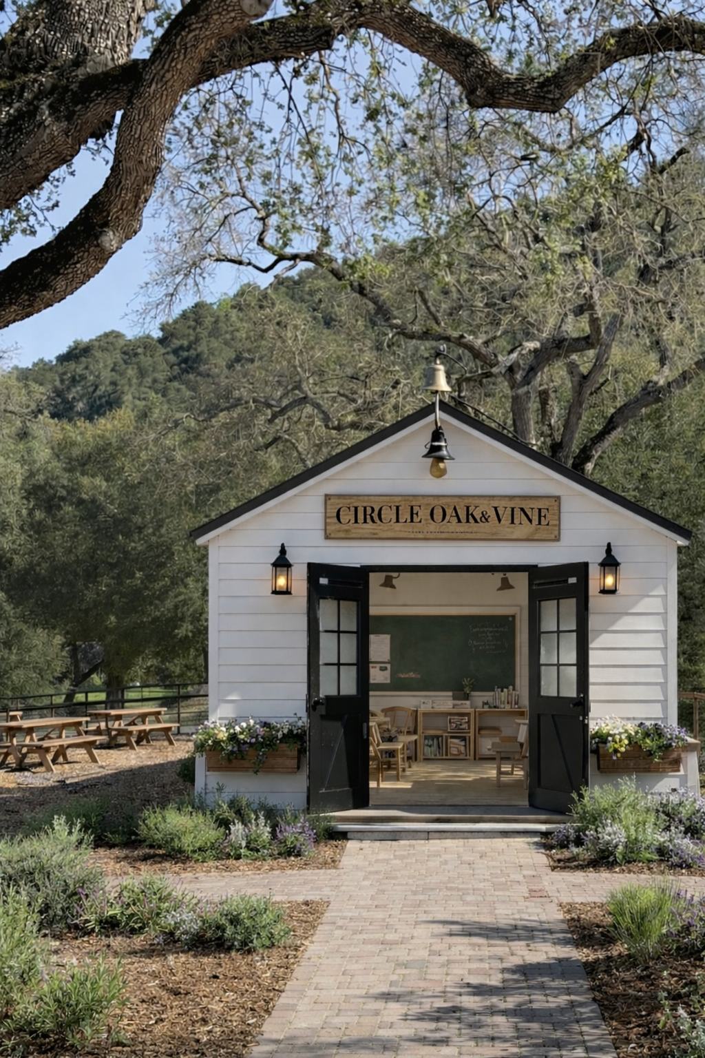 A small white building with black doors and a sign that reads 'Circle Oak & Vine' is set in a garden with flowering bushes. The building has an open double door, and inside there are chairs, a chalkboard, and a small table. The area is surrounded by trees and has a paved path leading to the entrance.