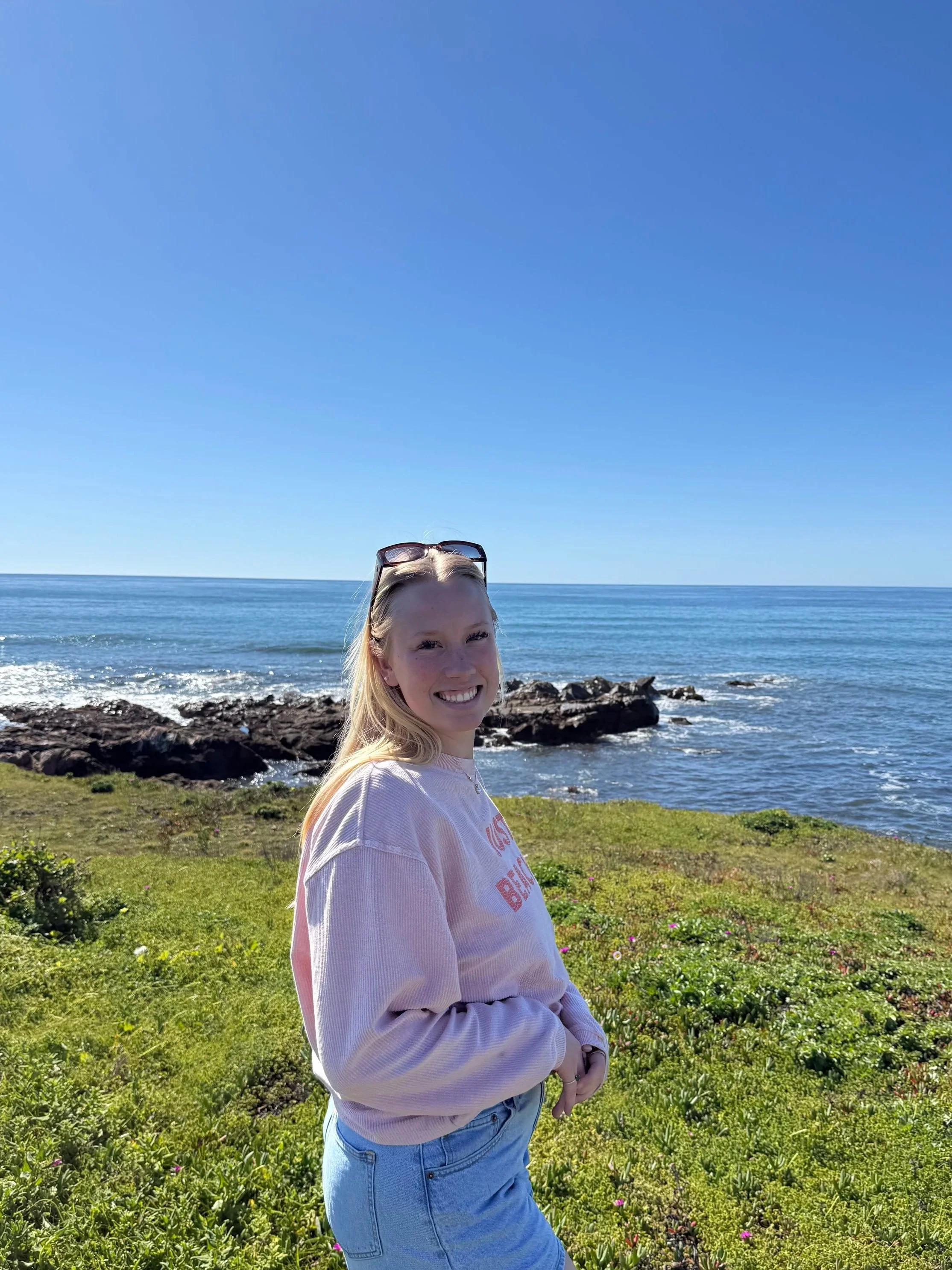 A young woman with long blonde hair smiling at the camera, wearing a pink sweatshirt and light blue jeans, standing on a grassy area near the ocean with rocks and the blue sky in the background.