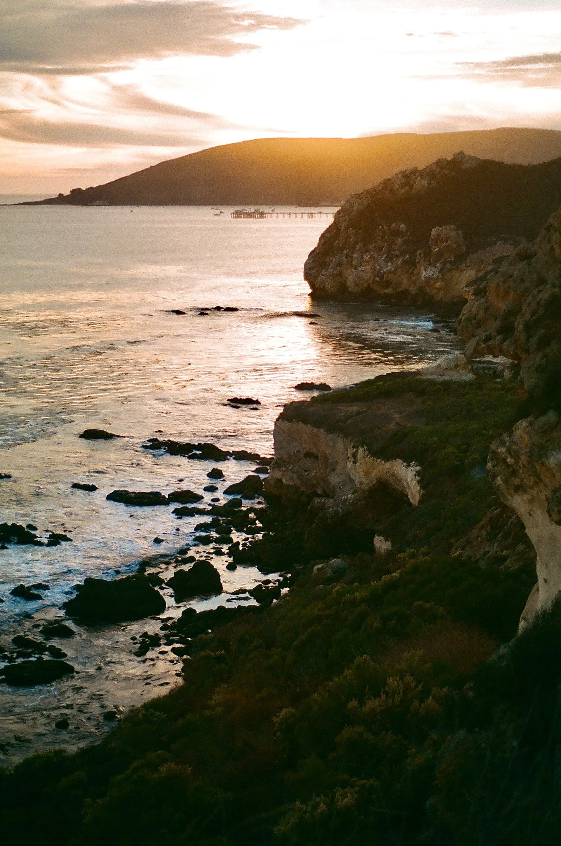 Sunset over a rocky coastline with the ocean and a distant hill or island in the background.