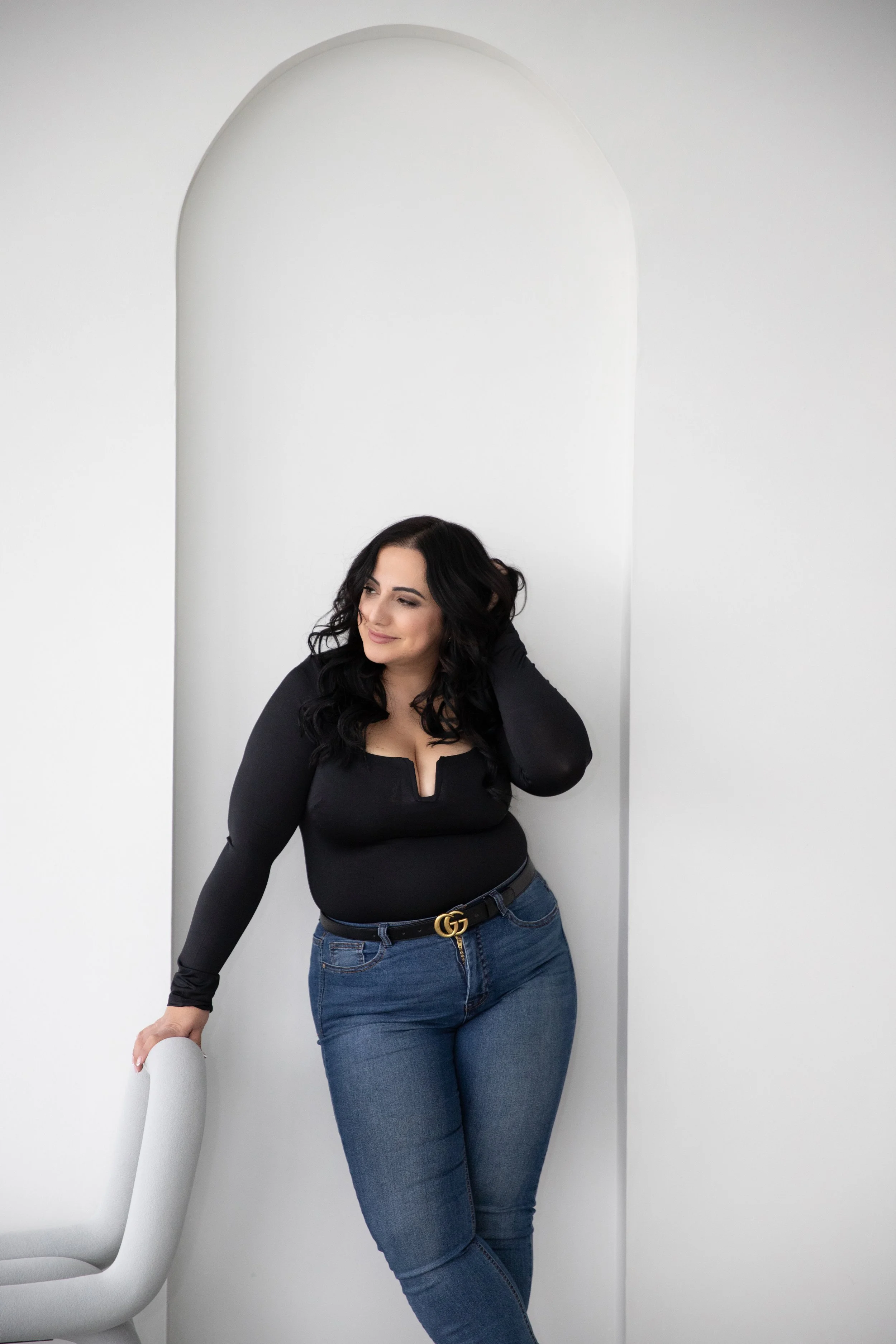 A woman with long black wavy hair, wearing a black top and blue jeans, is standing next to a black and white chair and smiling softly while looking to the side against a white wall with an arched cutout.
