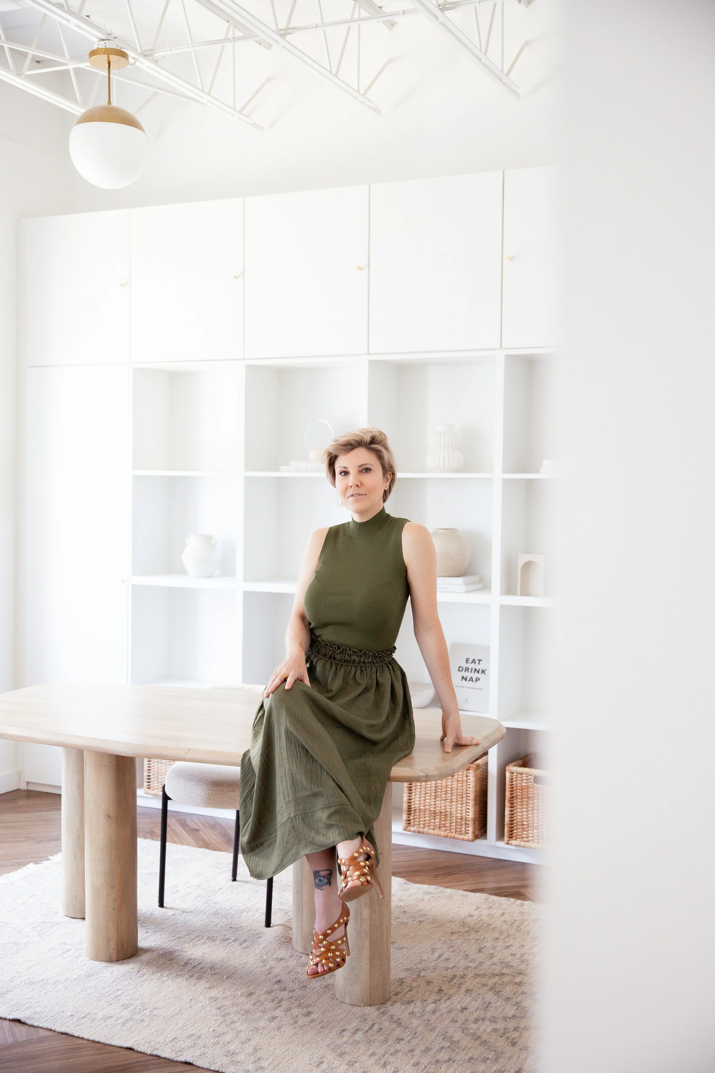 A woman with short blonde hair dressed in a sleeveless olive-green top and matching long skirt sitting on a wooden table in a bright, modern room with white walls and shelving, wicker baskets, and minimalist decor