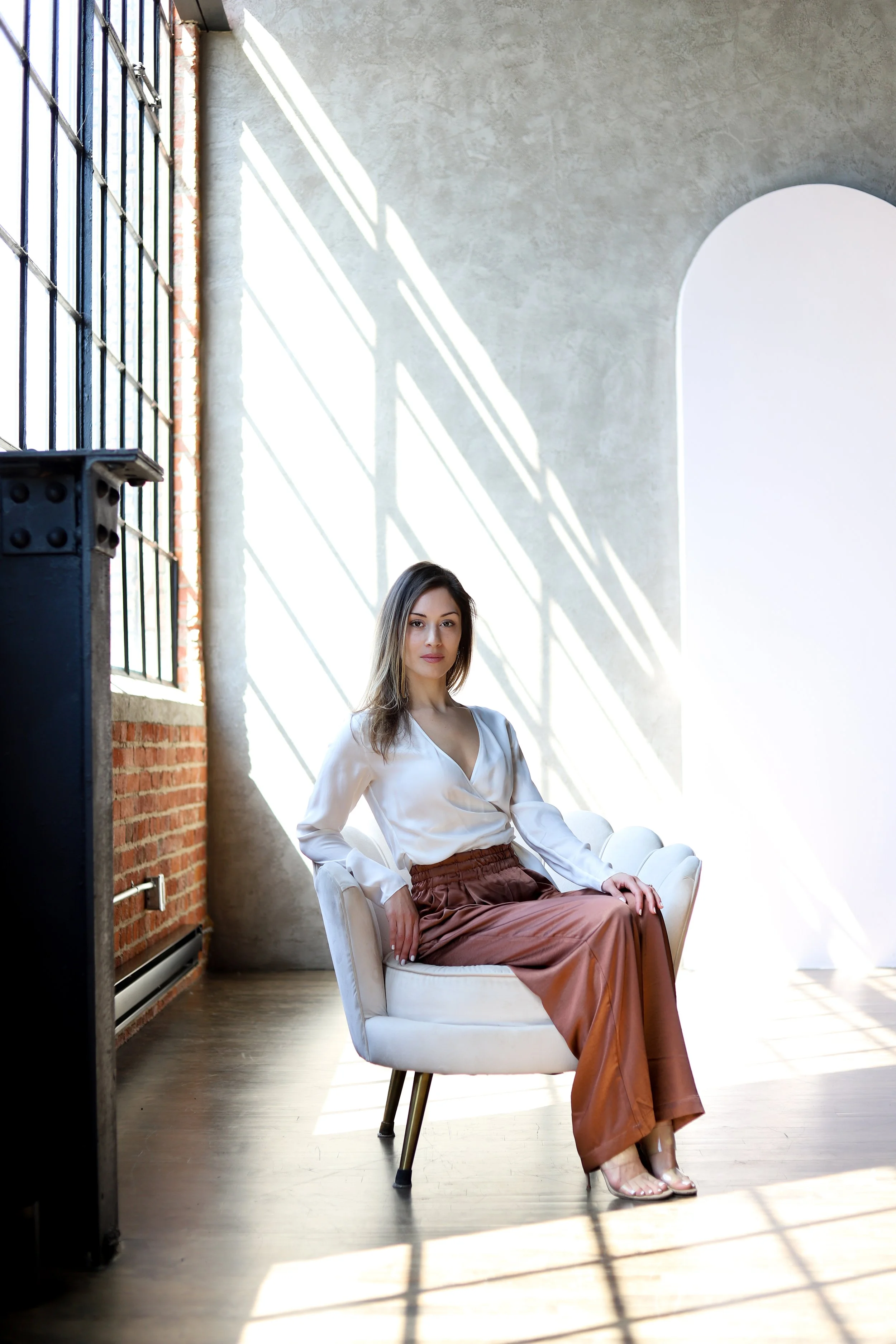 A woman with shoulder-length brown hair posing on a light-colored chair in a bright room with large windows. She is wearing a white blouse, brown wide-leg pants, and high-heeled sandals. Sunlight streaming through the windows creates shadows on the wall behind her.