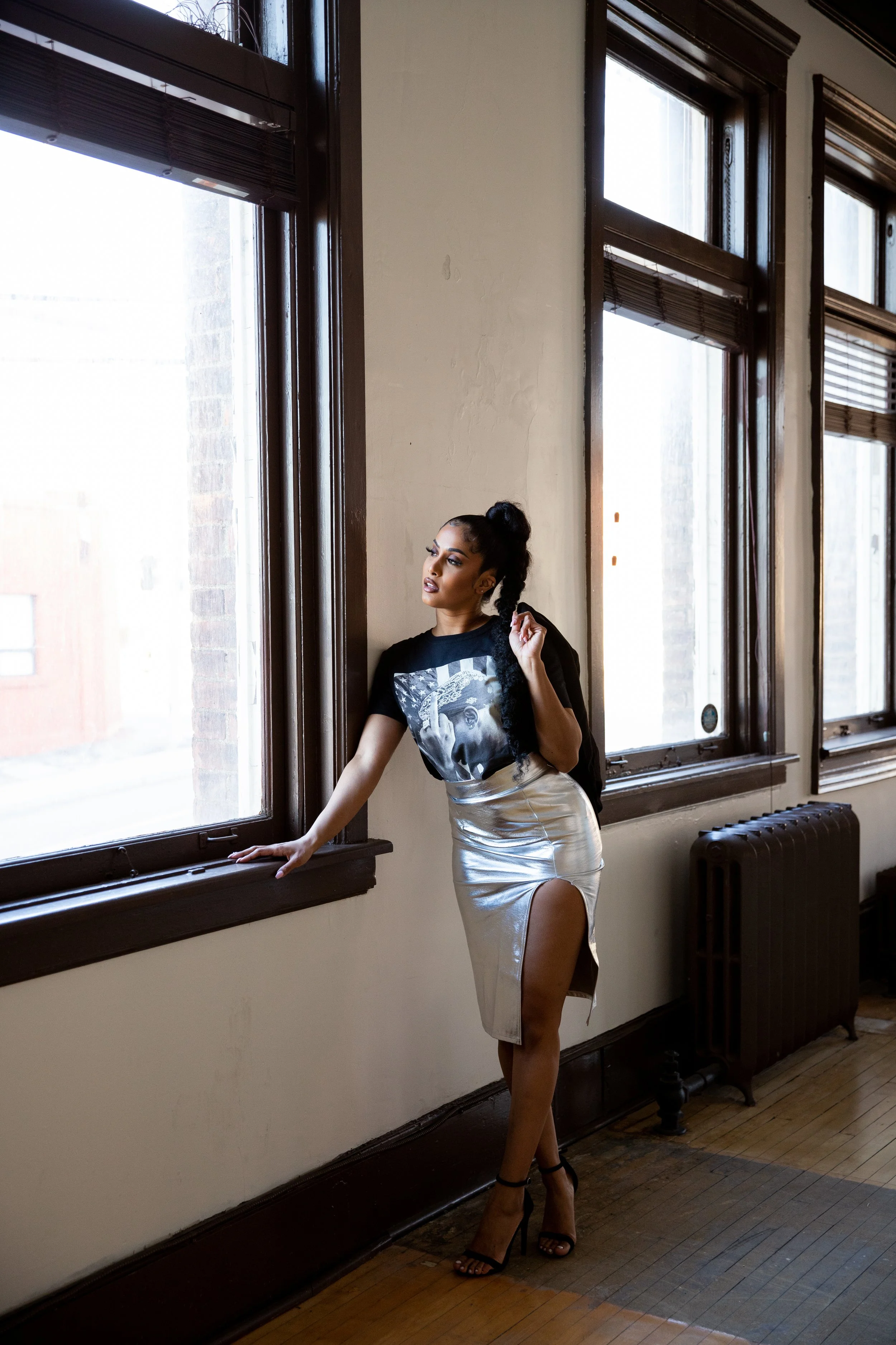 A woman with dark hair styled in a high bun, wearing a black T-shirt with a graphic print, a shiny silver skirt with a high slit, and black high heels, standing inside a room near large windows with wooden frames, looking out thoughtfully.