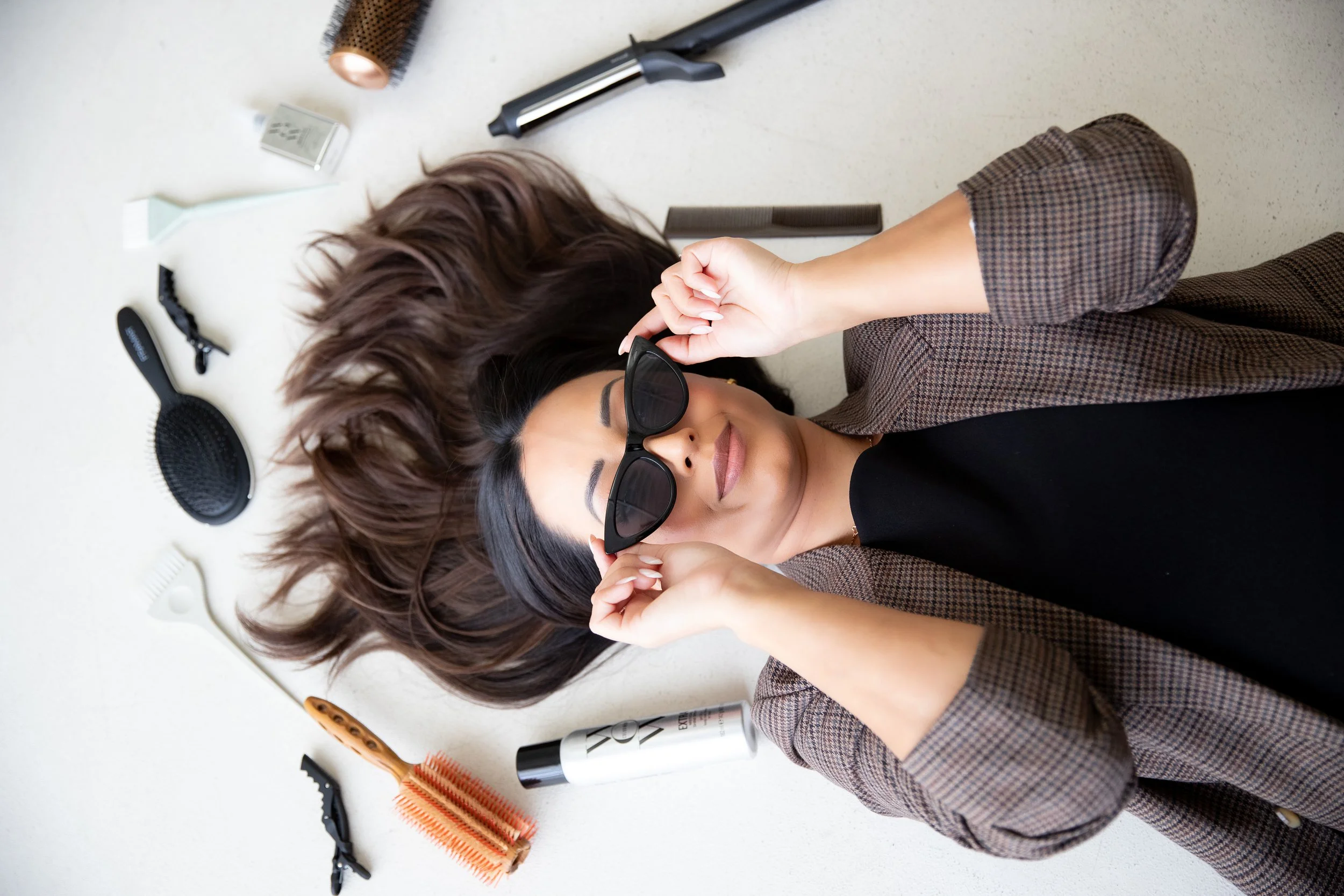 A woman with long brown hair lies on the floor surrounded by hair styling tools and products, holding sunglasses up to her face.