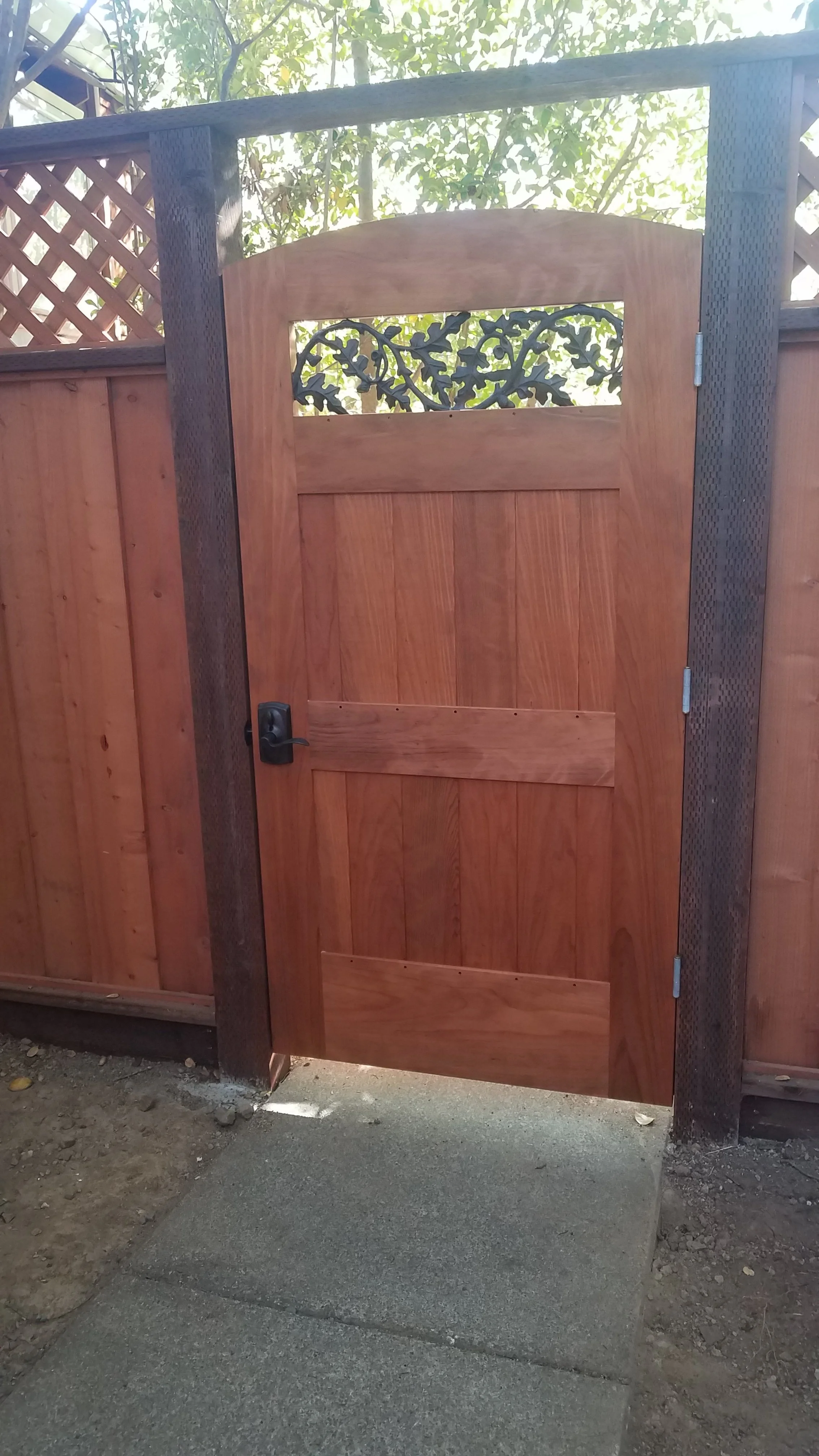 Wooden gate with metal accents next to a wooden fence, leading to an outdoor area with trees.