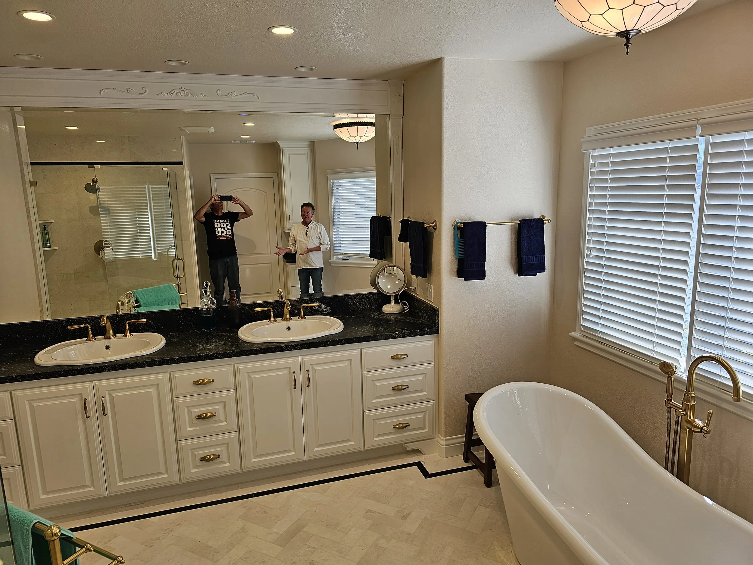 A spacious bathroom with a large mirror, double sinks with black countertops, a bathtub, and a walk-in shower. Two men are visible in the mirror, one taking a photo and the other talking.