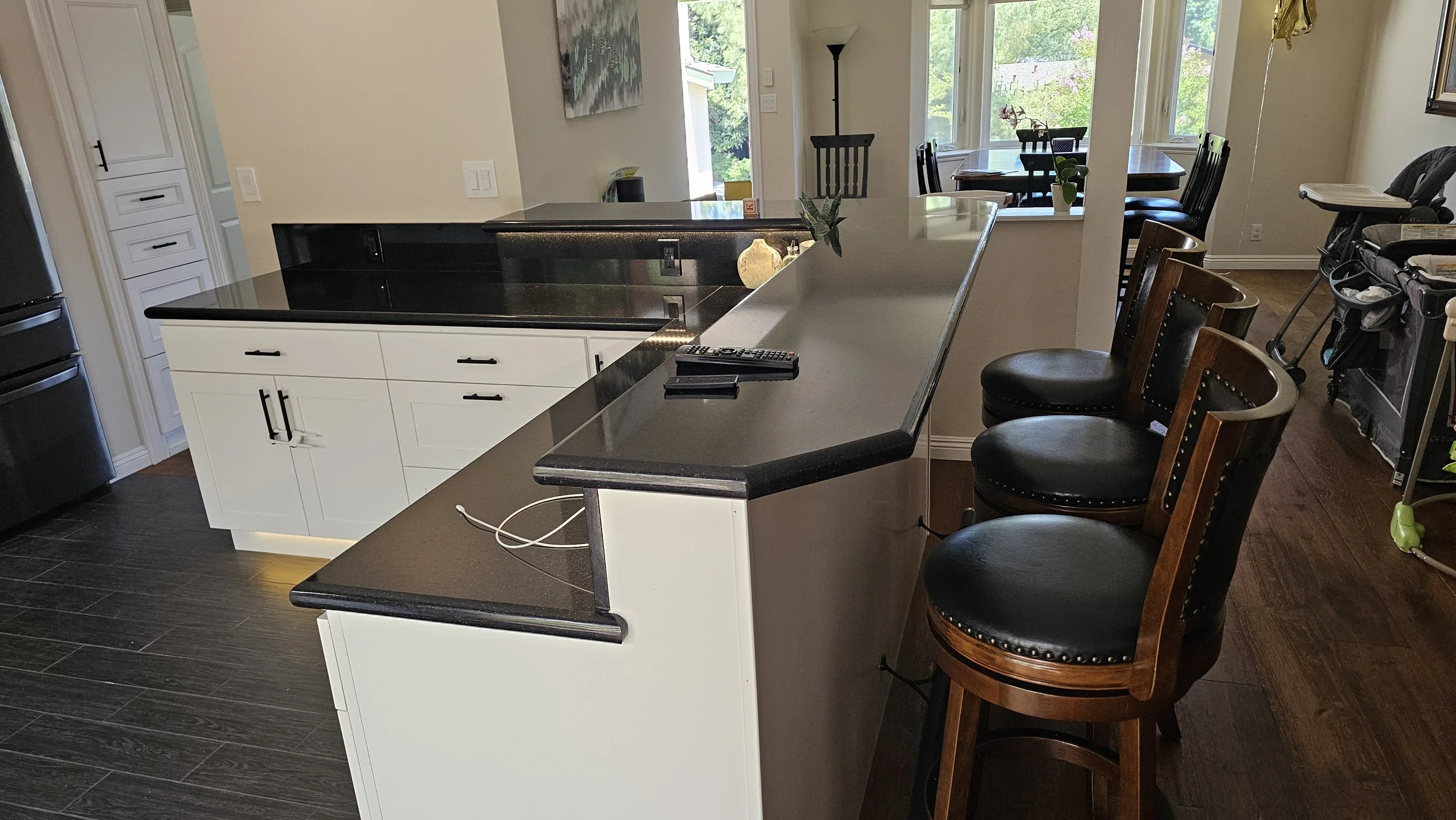 Kitchen with white cabinets and black countertops, black and dark wood barstools, dining table with chairs, windows showing green trees outside, and a baby high chair.