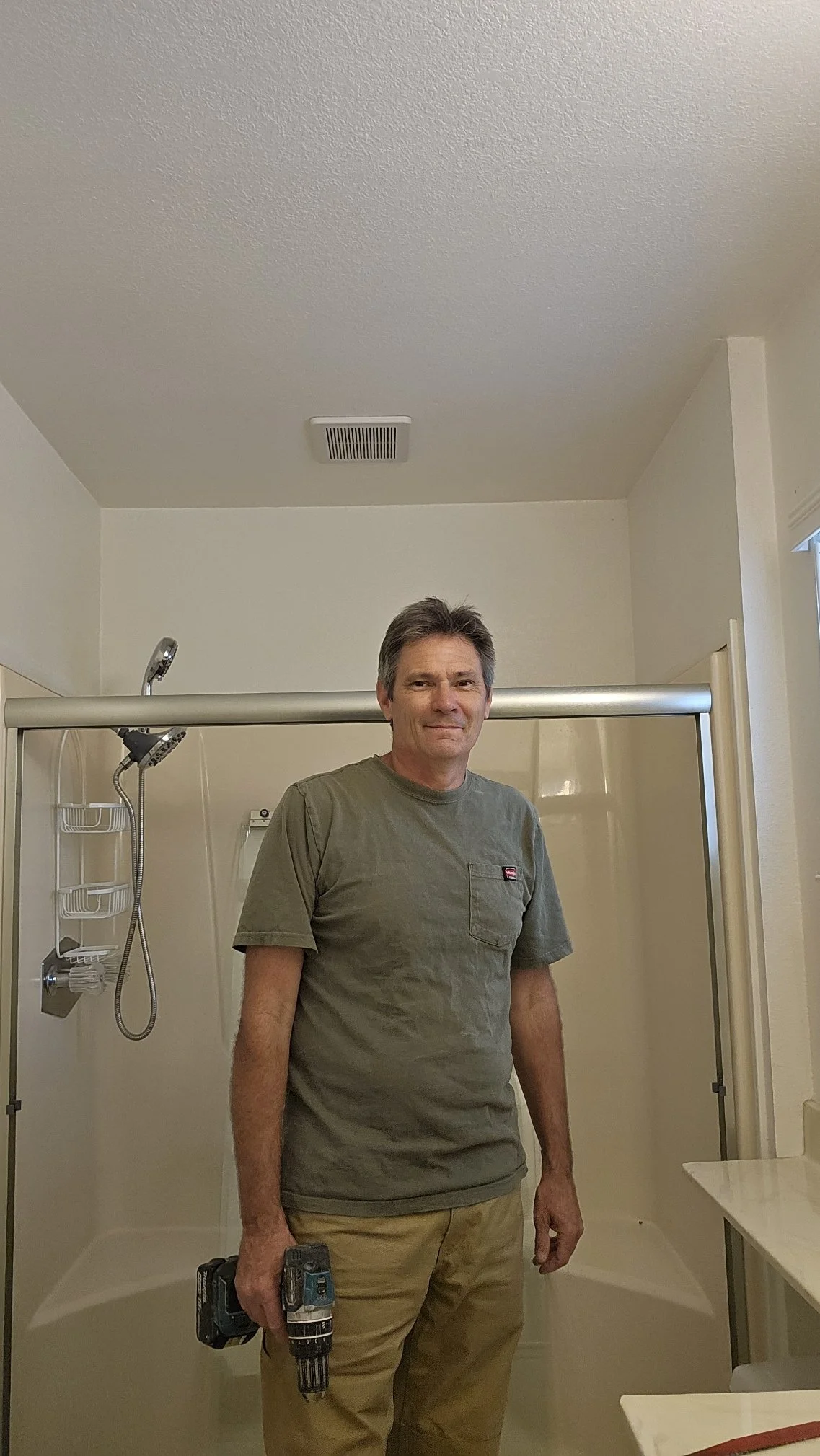 A man standing in a shower area holding a cordless drill, with the showerhead and bathroom fixtures visible in the background.