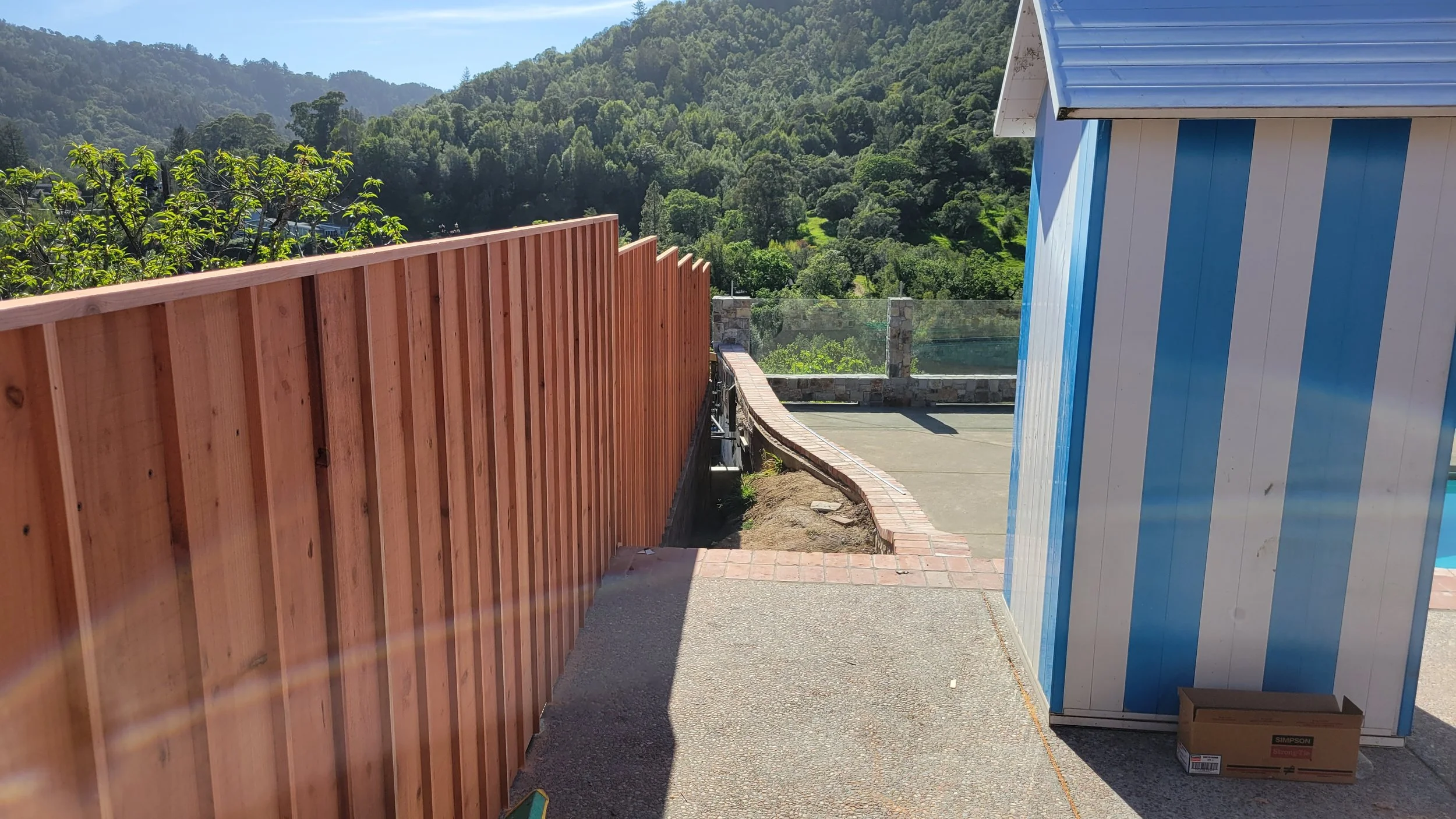 Outdoor area with a wooden fence on the left, a blue and white striped shed on the right, and a mountain landscape in the background under a clear sky.