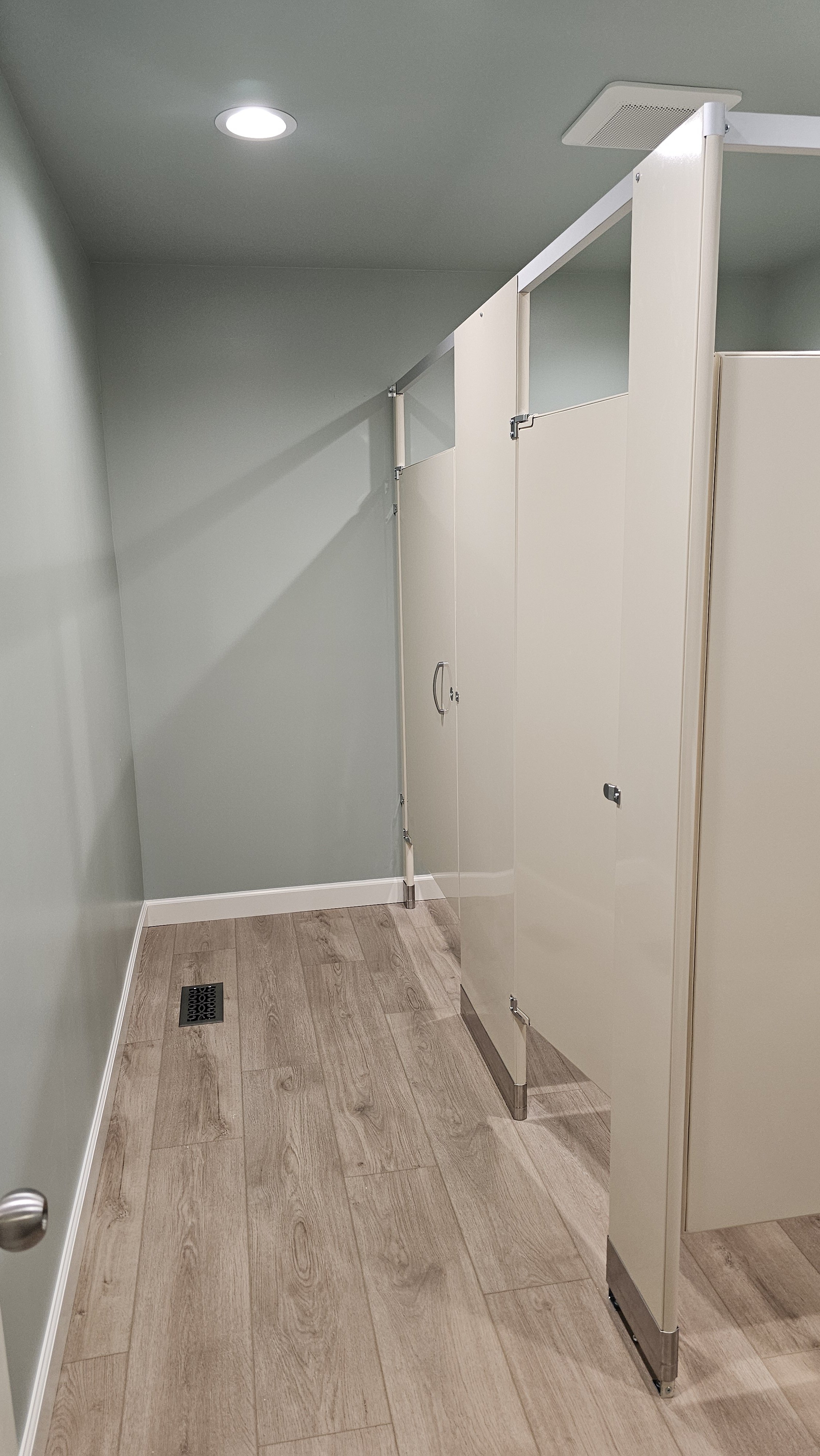 Empty public restroom stall with beige partition doors, wood-look flooring, gray walls, and recessed ceiling light.