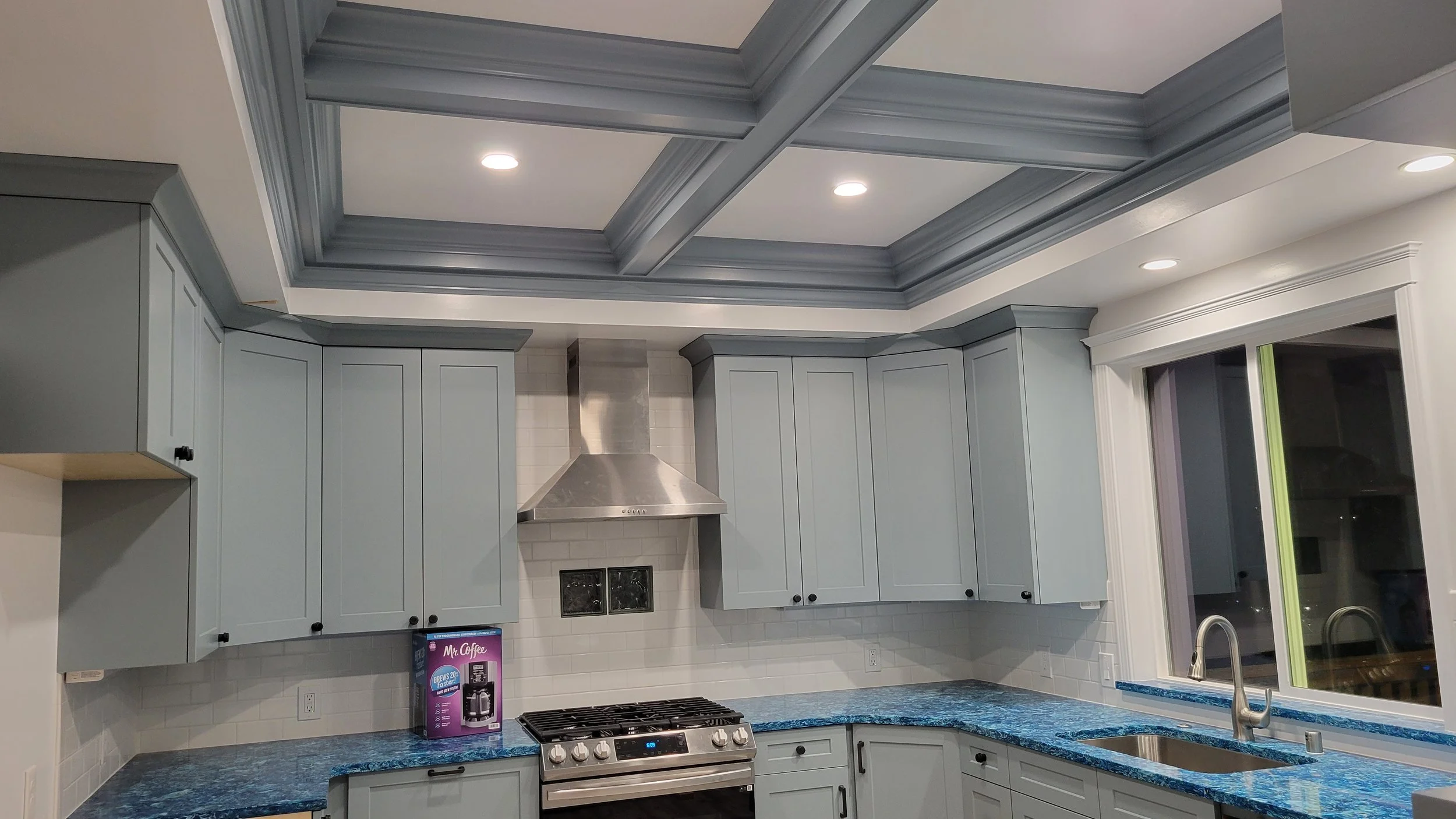 Modern kitchen with light blue cabinets, a blue granite countertop, stainless steel stove and range hood, white tiled backsplash, a large window above the sink, and recessed lighting in the ceiling.