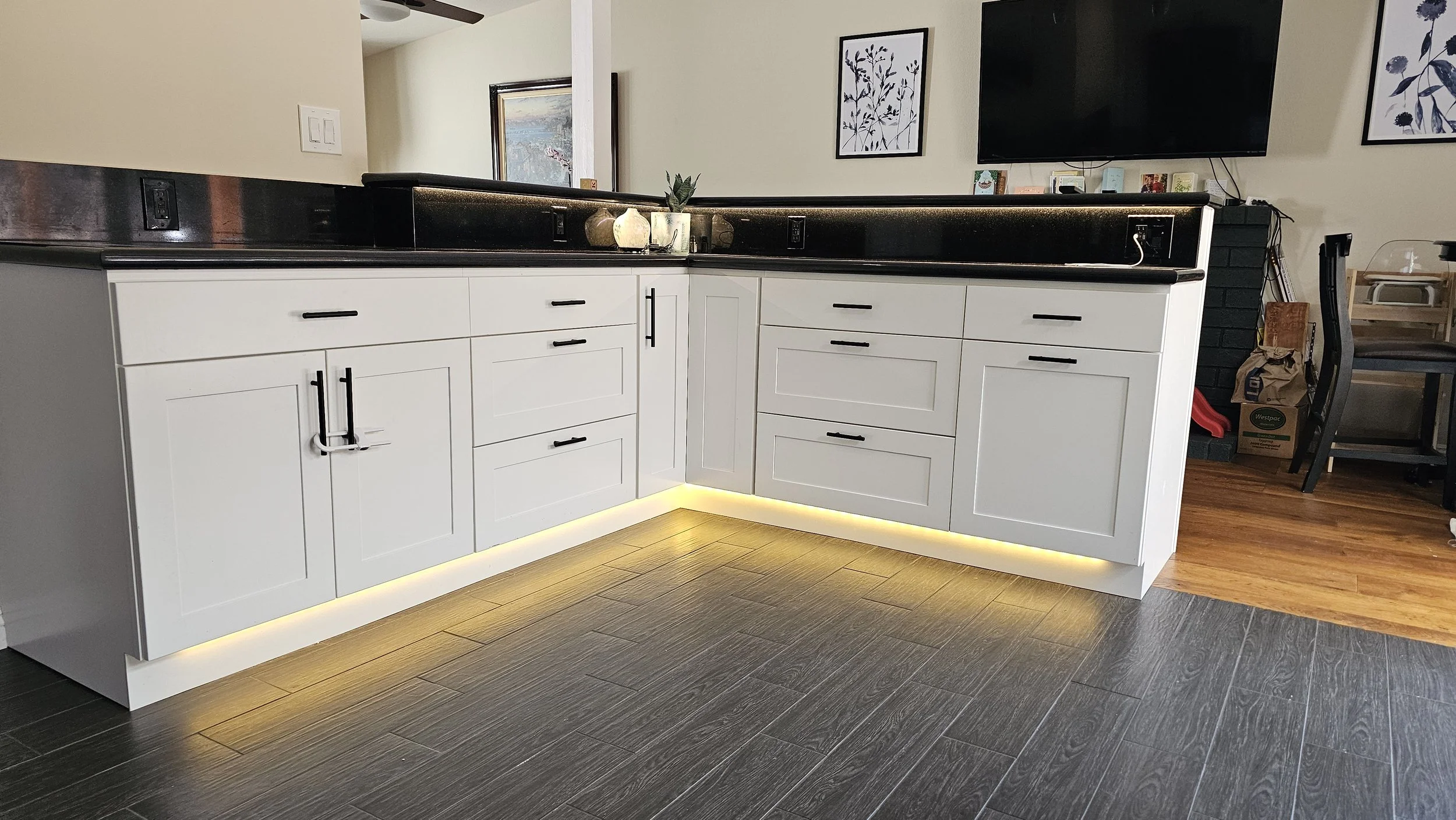 Modern kitchen with white cabinets, black handles, black countertop, and LED lighting near the floor. There are decorative objects on the counter, a wall-mounted TV, and framed artwork on the wall.