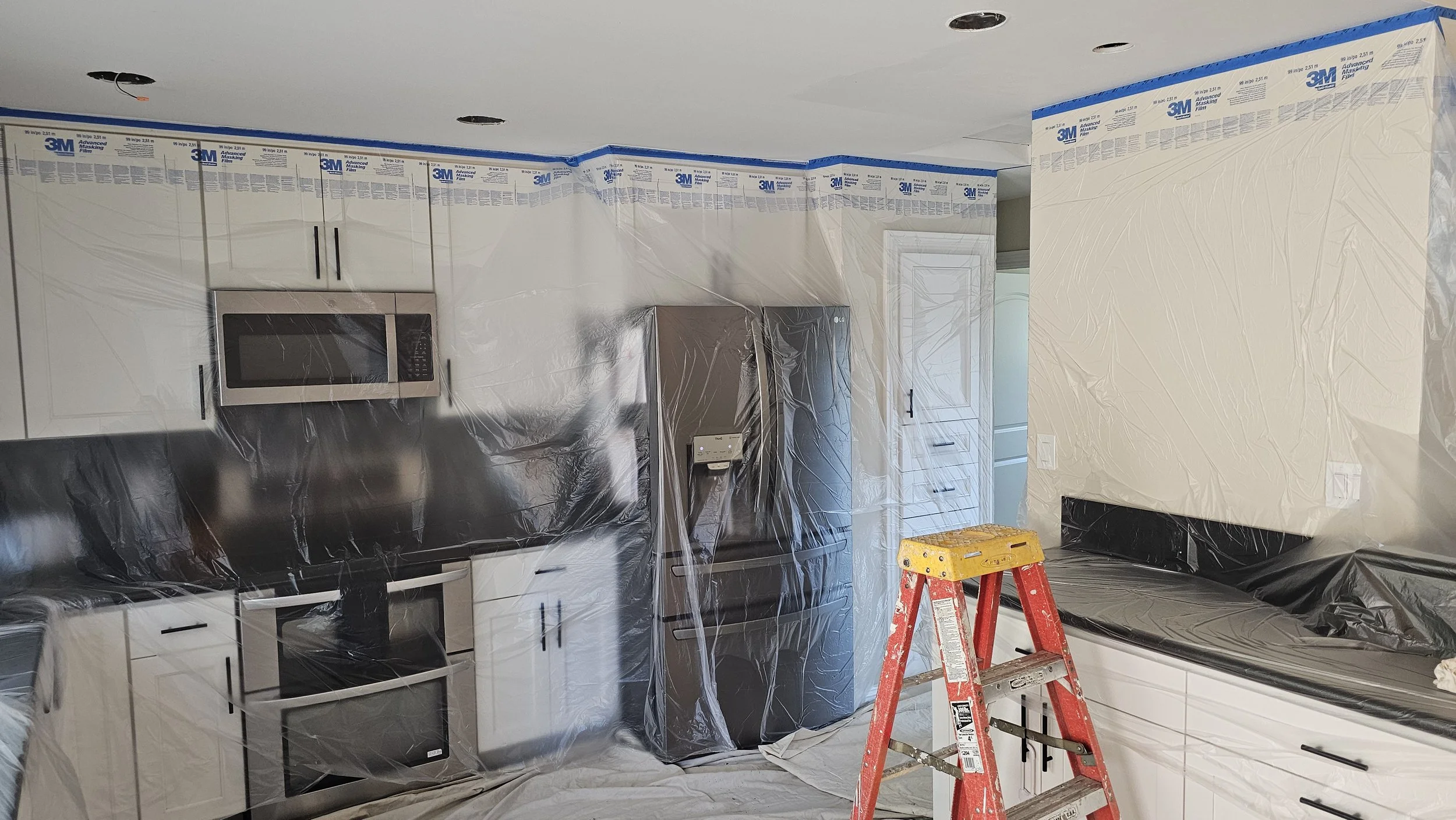 Kitchen under renovation with plastic sheeting covering cabinets, countertops, and appliances; a ladder in the center; some wall outlets and lights visible; ceiling is unfinished with holes for lighting.