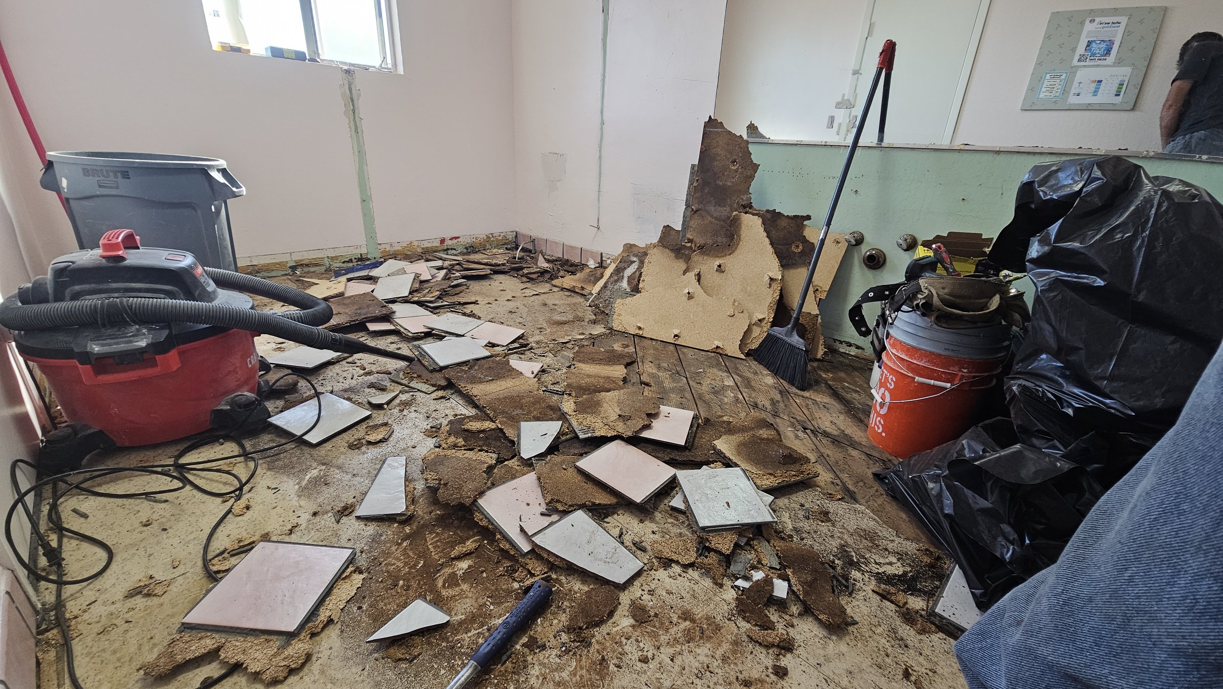 Room undergoing renovation with torn drywall and broken tiles scattered on the floor, construction tools including a wet vacuum, a broom, and a hammer visible, along with trash bags and cleaning supplies.