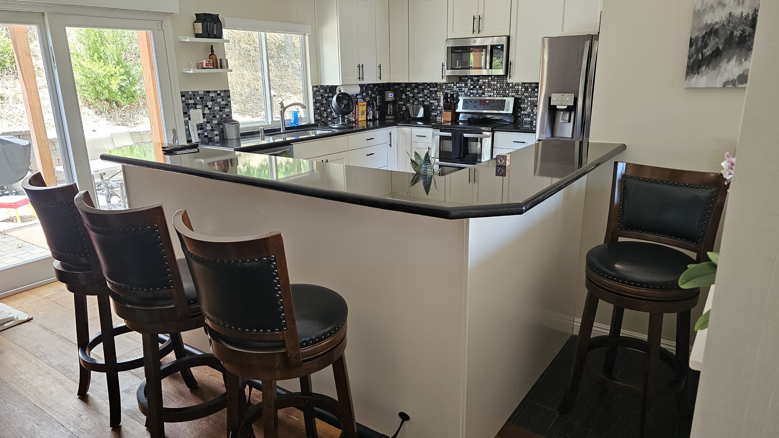 Kitchen with white cabinets, black backsplash, stainless steel appliances, and a black countertop bar with seating. There are three wooden bar stools with black cushions around the bar, and natural light coming through large sliding glass doors.