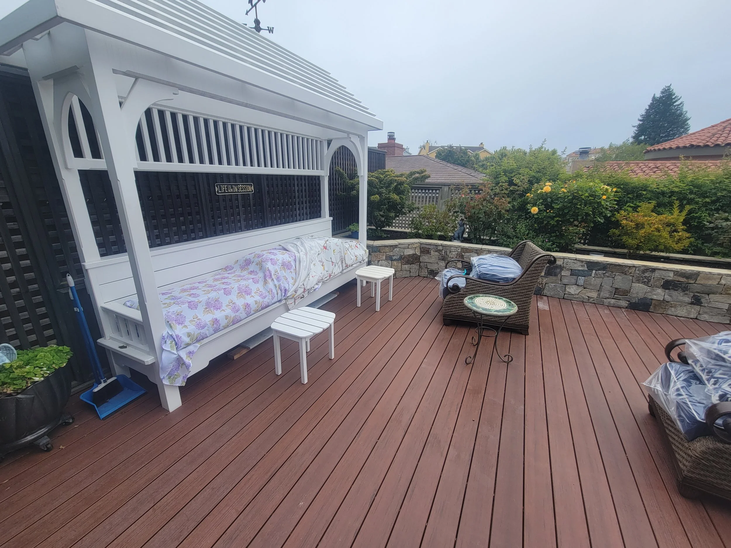 A wooden deck outdoor patio with furniture, including a swing with a canopy, small side tables, a wicker chair, and a stone retaining wall with plants and flowers in the background, under a cloudy sky.