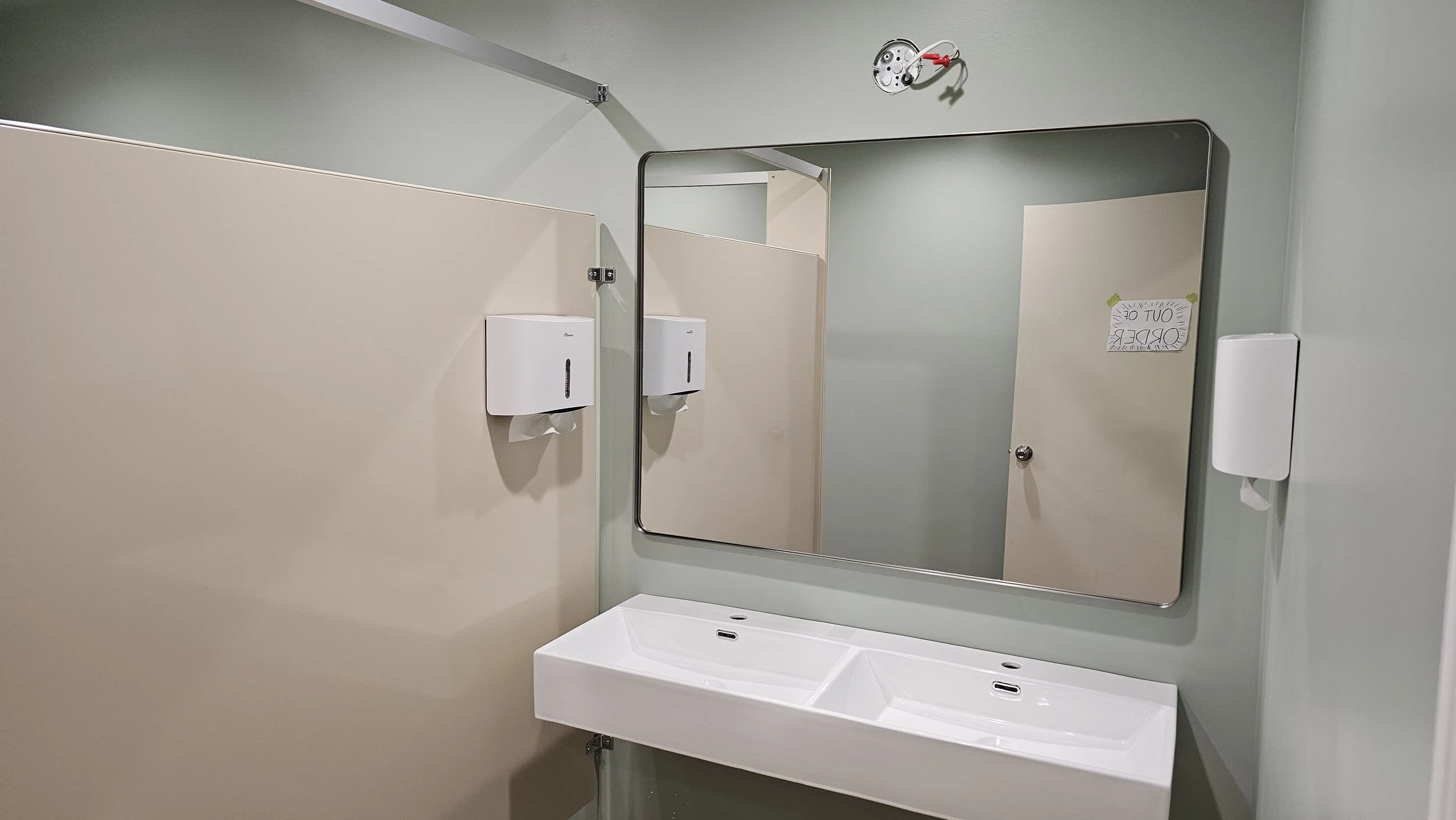 Public restroom with a large mirror, white sink, soap dispensers, and a sign that says 'Out of Order' taped to the door.