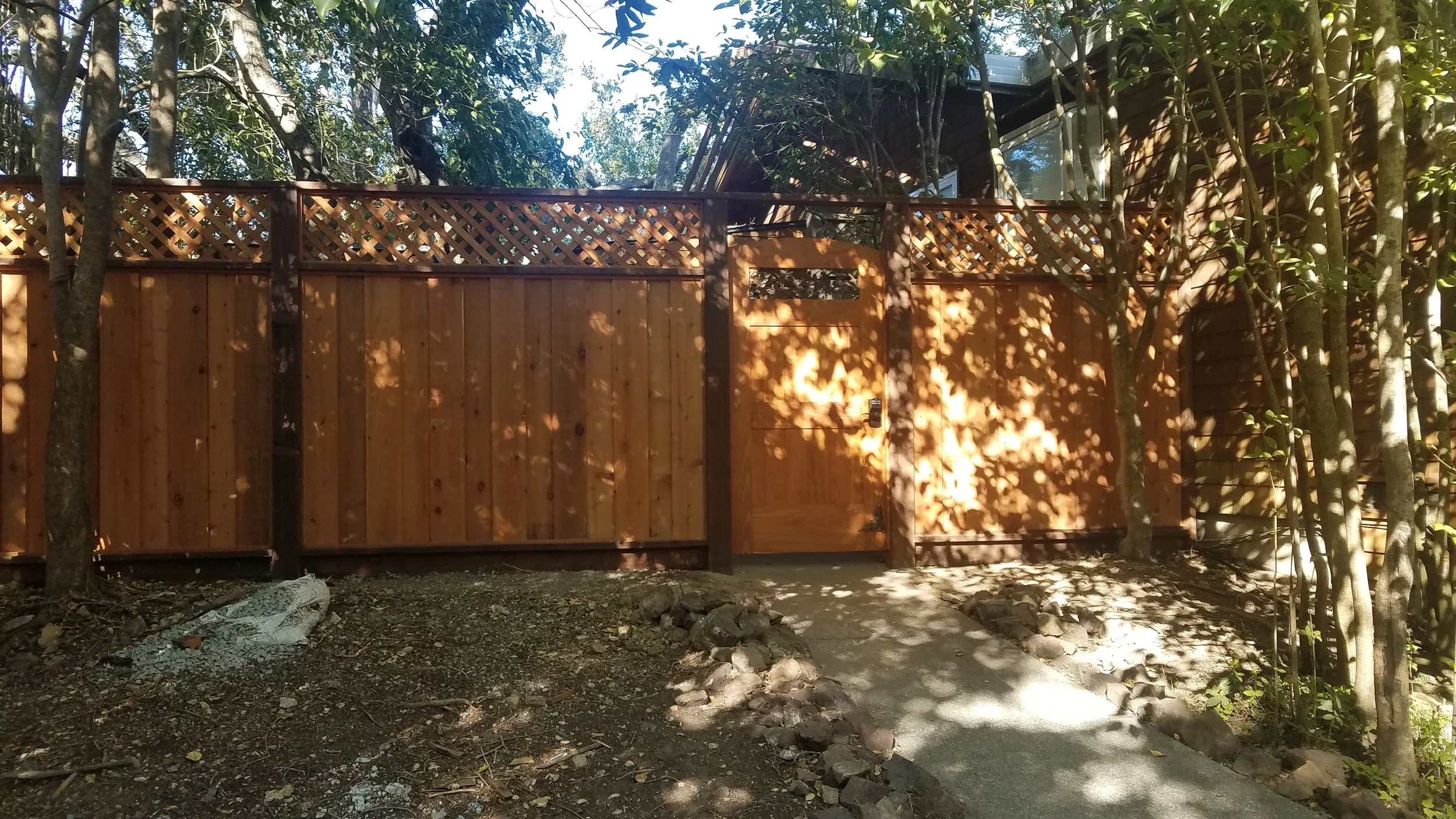 Wooden fence with gate surrounded by trees and shadows on the ground and fence, with a pathway leading to the gate.