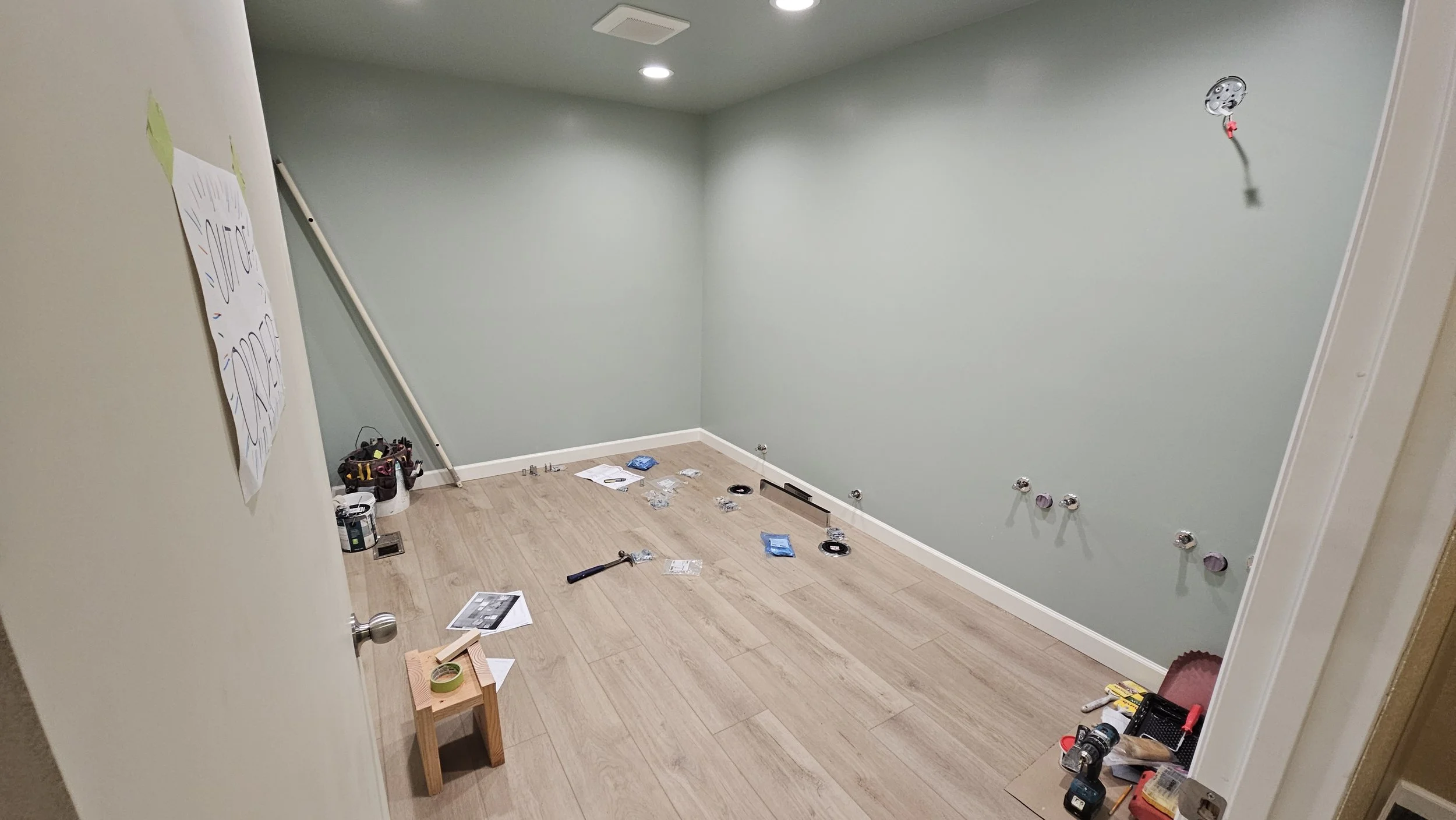 Room with light green walls and wooden floor, containing tools, hardware parts, and wiring for electrical work, with a ladder leaning against the left wall.