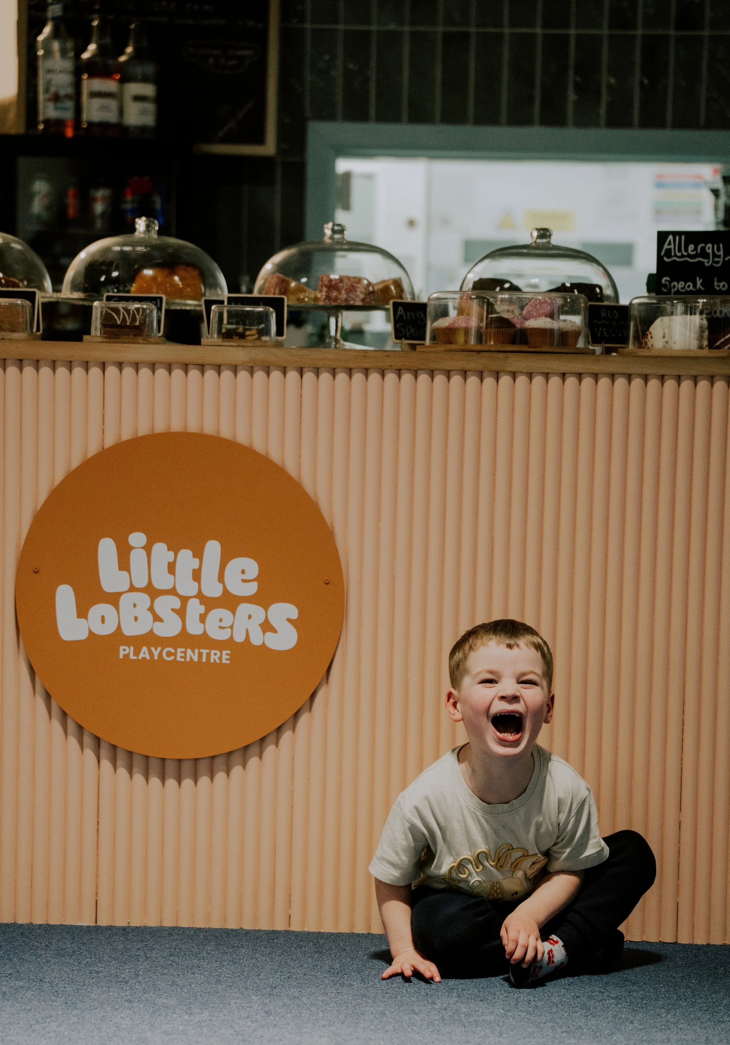A young boy crouching and laughing with his mouth open, in front of a play center sign that reads 'Little Lobsters Play Centre.' The background includes a colorful wooden wall, and the boy is wearing a gray T-shirt with a graphic and dark pants.