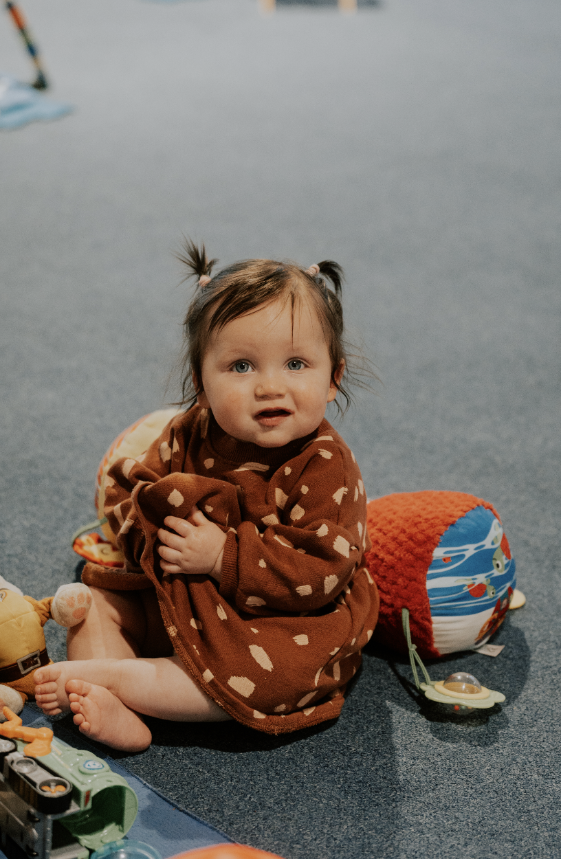 A young girl with brown hair in pigtails sitting on a carpeted floor, wearing a brown patterned dress, surrounded by toys, with a curious expression.
