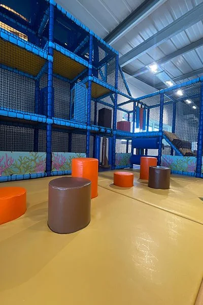Indoor playground with colorful padded structures, including orange and brown cylindrical seats, and a multi-level climbing structure with blue netting and yellow platforms inside a large, airy space.
