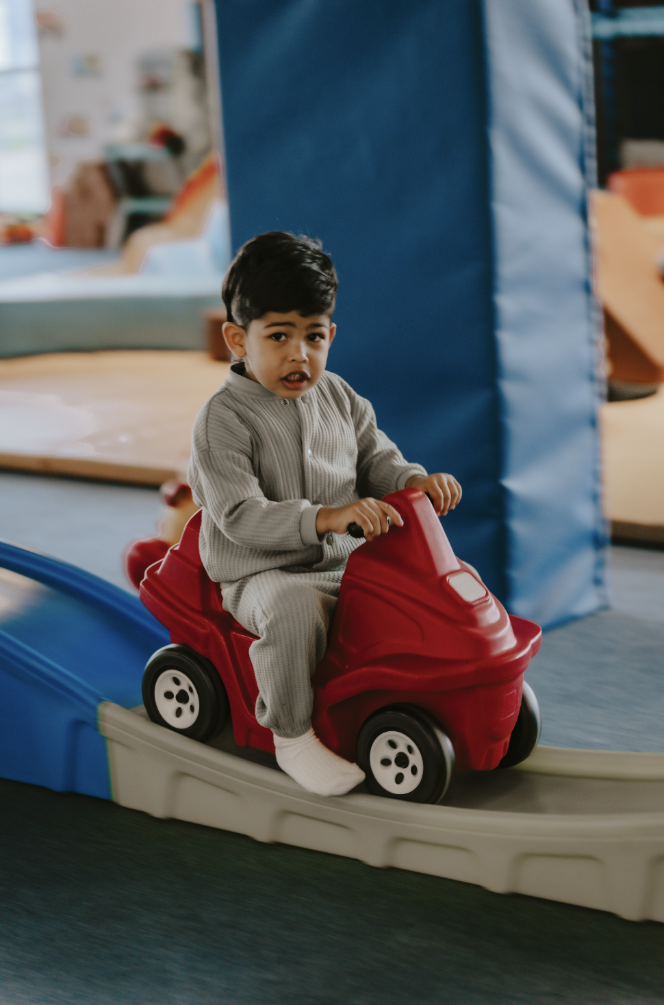 A young boy in gray pajamas riding a red toy car on an indoor track.