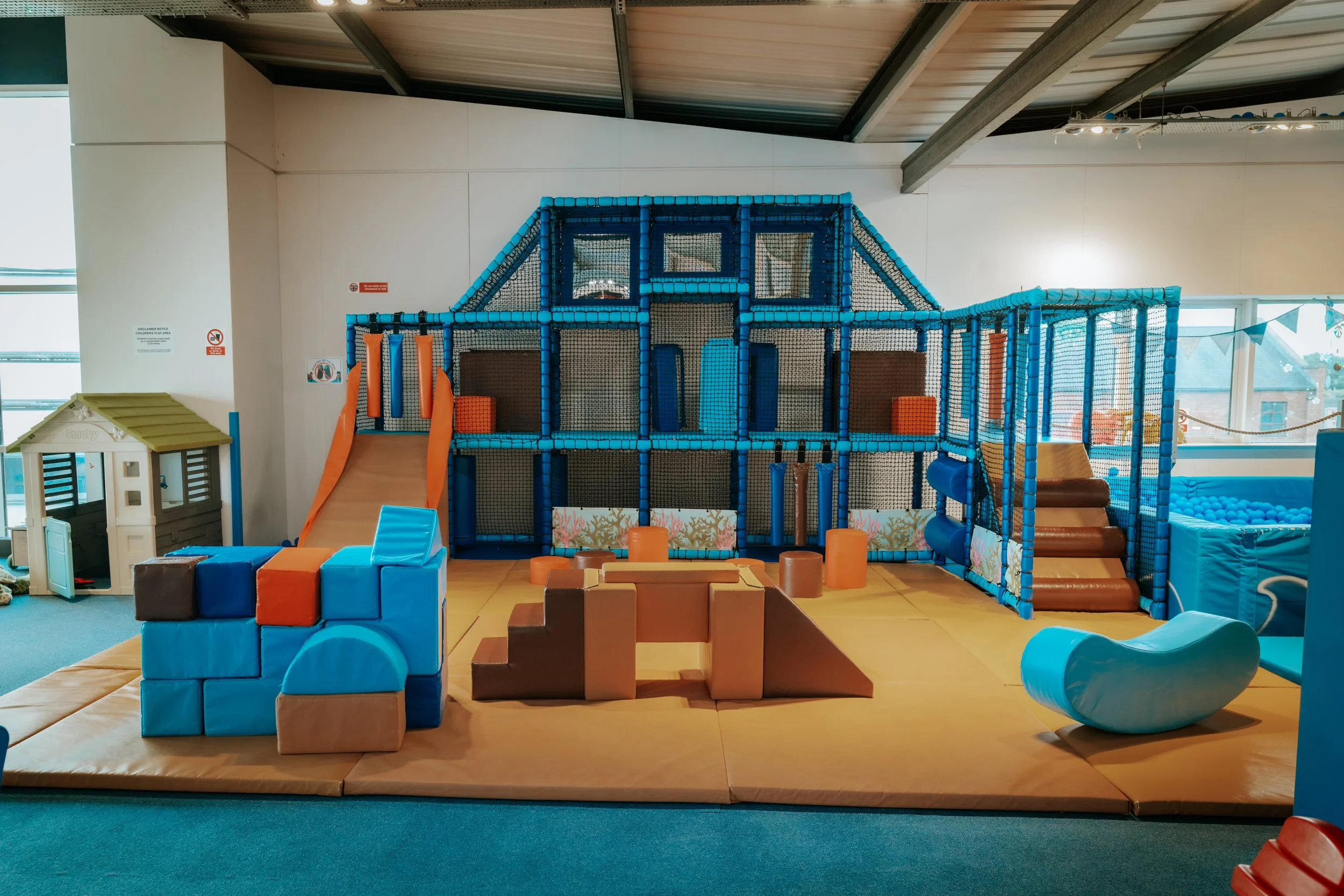 Indoor children's play area with a blue and orange climbing structure, slides, and soft foam mats.