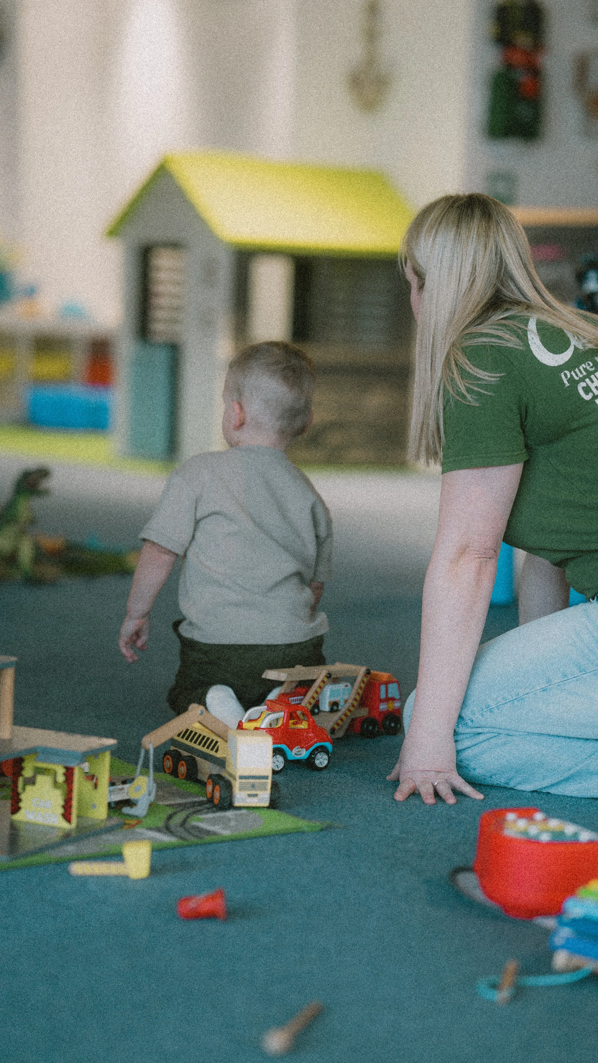 A woman and a young boy playing with toy vehicles on a colorful play mat in a play area.