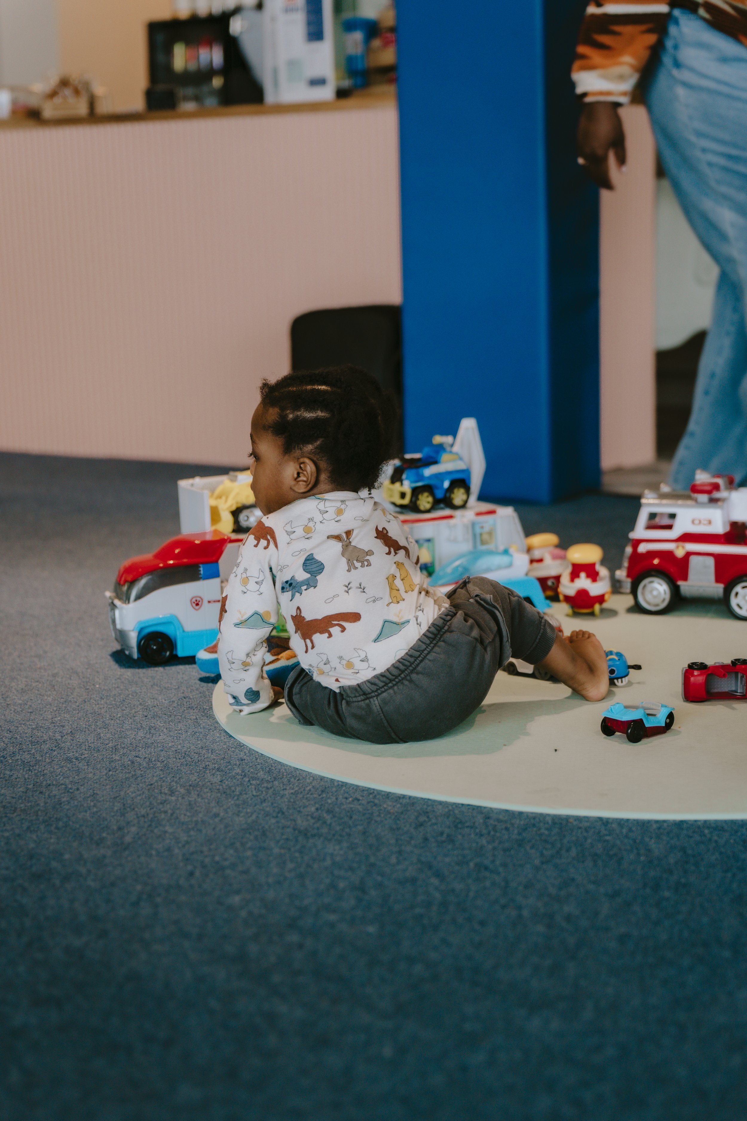 Child with braided hair sitting on the floor next to toy vehicles and figurines, with partial adult visible on the right, in a room with blue and beige walls and a carpeted floor.