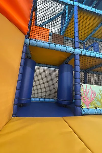 View of an indoor playground with colorful padded climbing structures, netted barriers, and soft flooring.