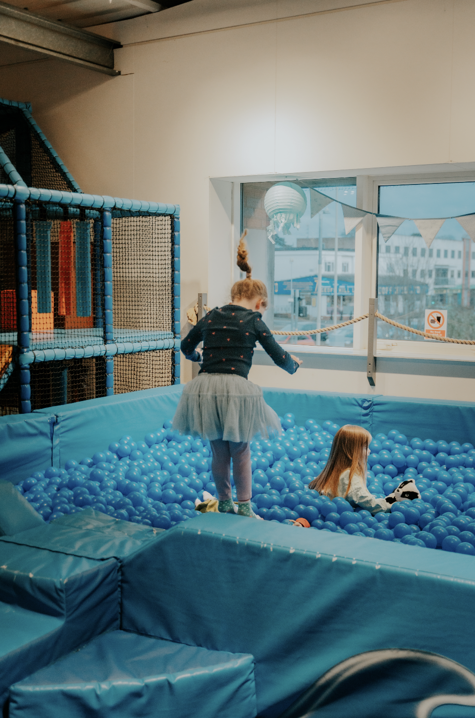 Two children playing in an indoor ball pit, one girl standing and the other sitting, surrounded by blue plastic balls.